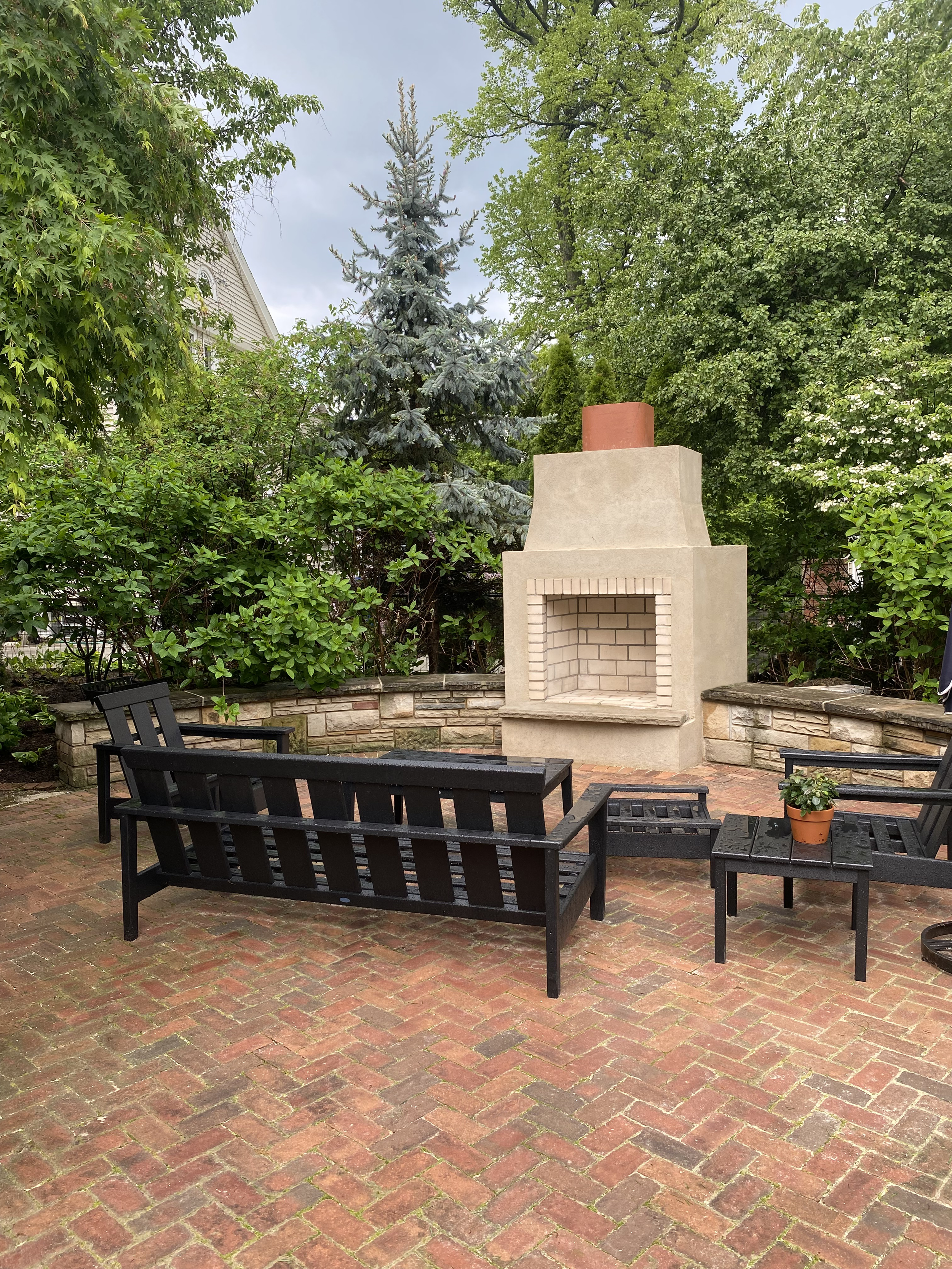 Backyard patio with brick flooring, black wooden benches, a small black table with a potted plant, and a beige outdoor fireplace with a chimney, surrounded by green bushes and trees.