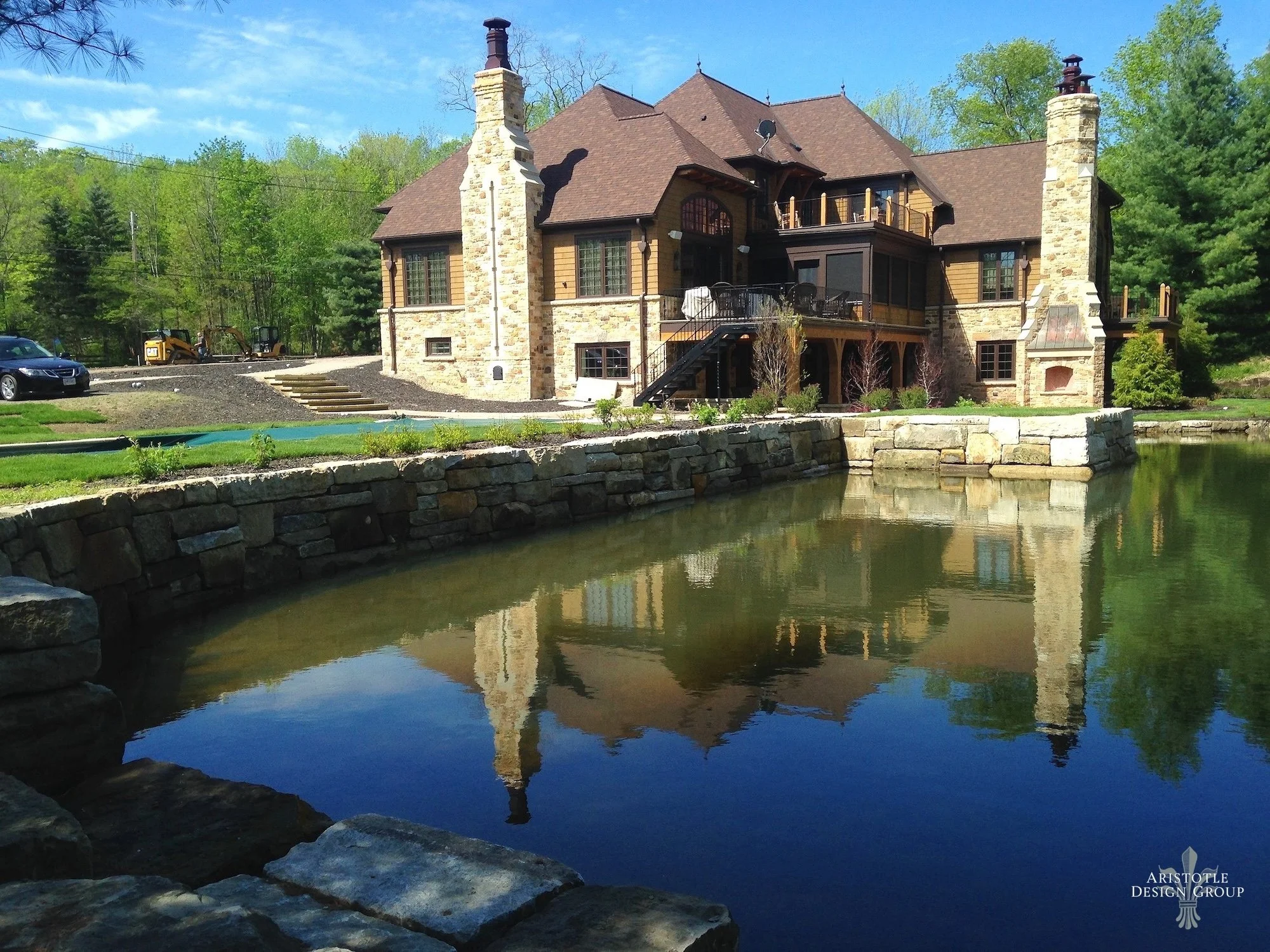 Large house with a stone and wood exterior, multiple chimneys, and a balcony, situated by a pond reflecting the house and trees, with a clear blue sky and green trees in the background.