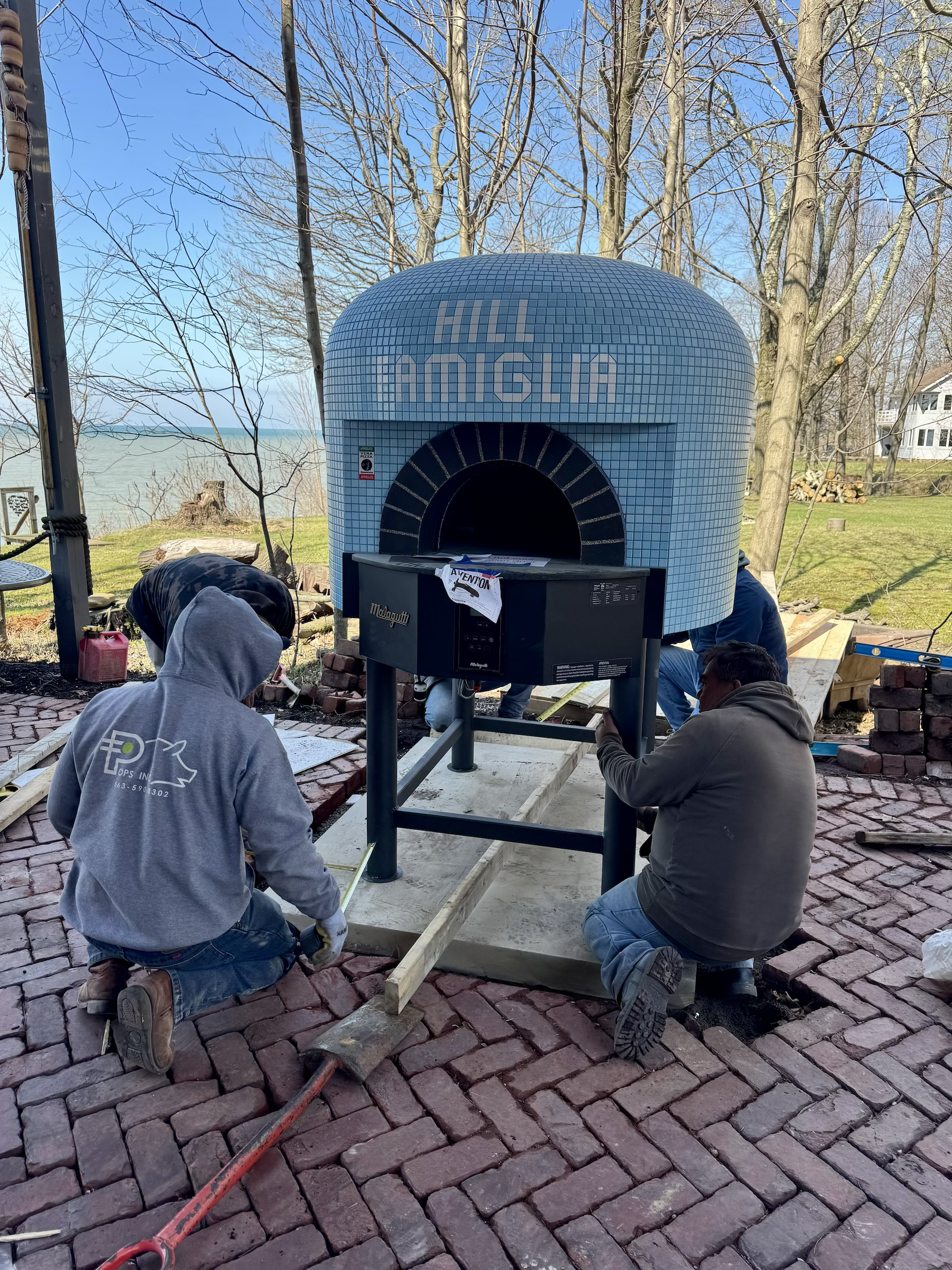 People installing a wood-fired pizza oven on a brick patio with a scenic lakeside view and leafless trees in the background.