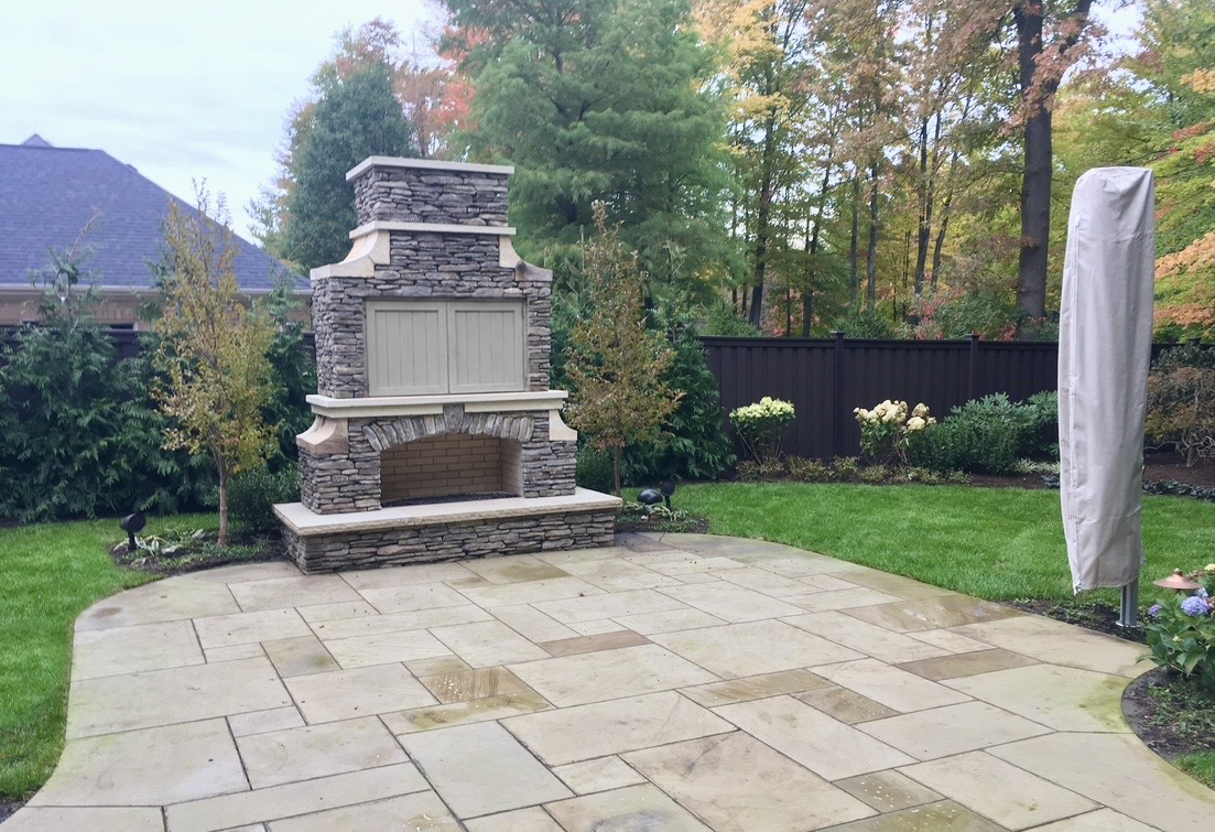 Backyard with a stone outdoor fireplace on a patio, surrounded by trees, bushes, and a black fence.