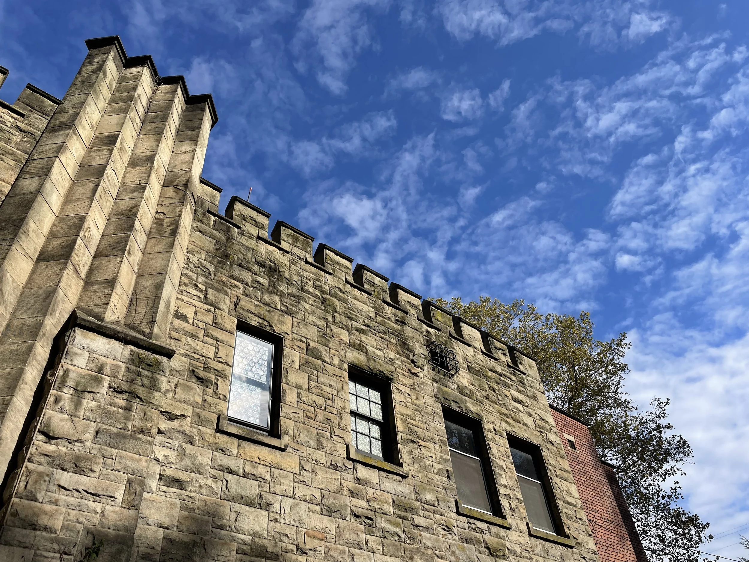 A stone building with several windows, topped with battlements, against a bright blue sky with scattered clouds, and a tree partially visible on the right.