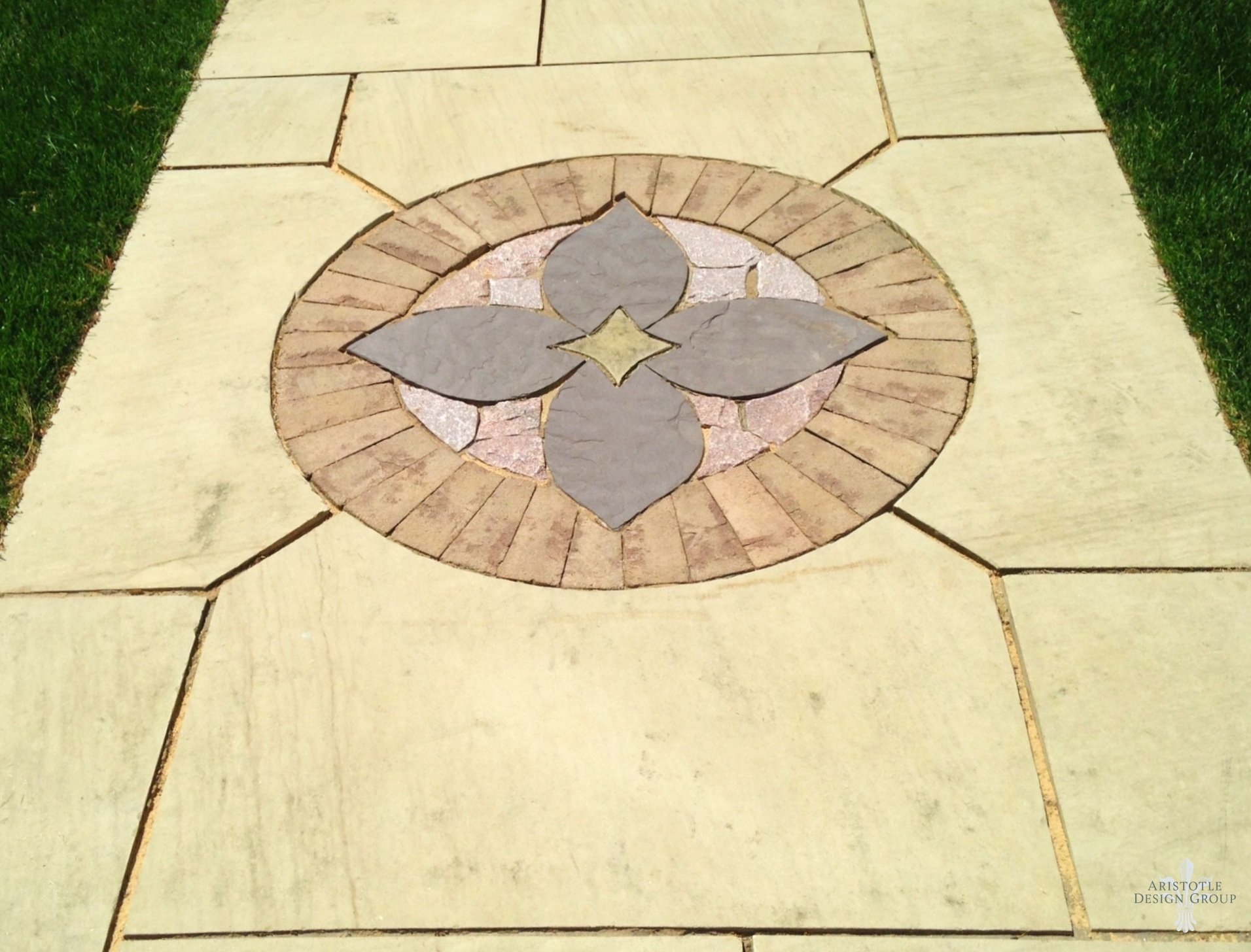 A decorative stone sidewalk with a circular pattern of colored stones in a floral design, surrounded by large rectangular paving stones and green grass.