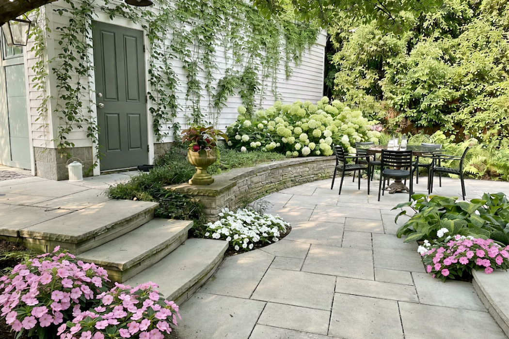 A backyard patio with a table and six chairs, surrounded by lush green plants and colorful flowers, including pink, white, and green hydrangeas, adjacent to a white house with climbing vines on the wall.