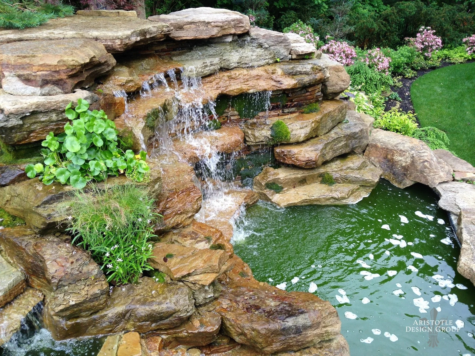 A small waterfall flowing over rocks into a pond surrounded by lush green plants and flowers in a garden.