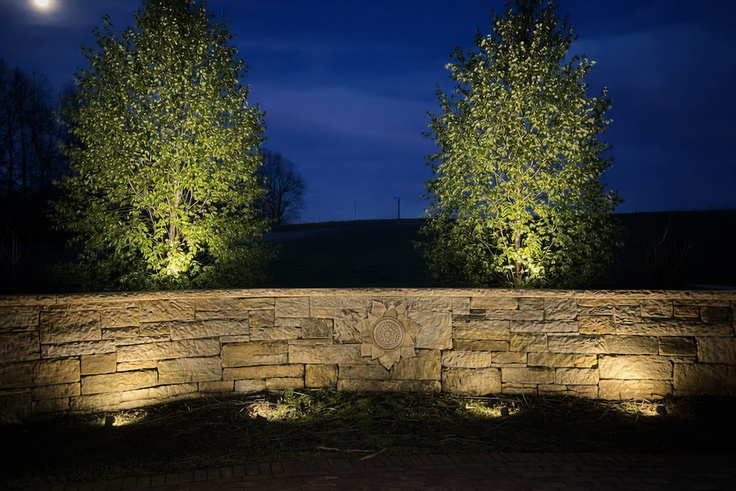 Nighttime scene with two illuminated trees behind a stone wall and a carved emblem in the center of the wall.