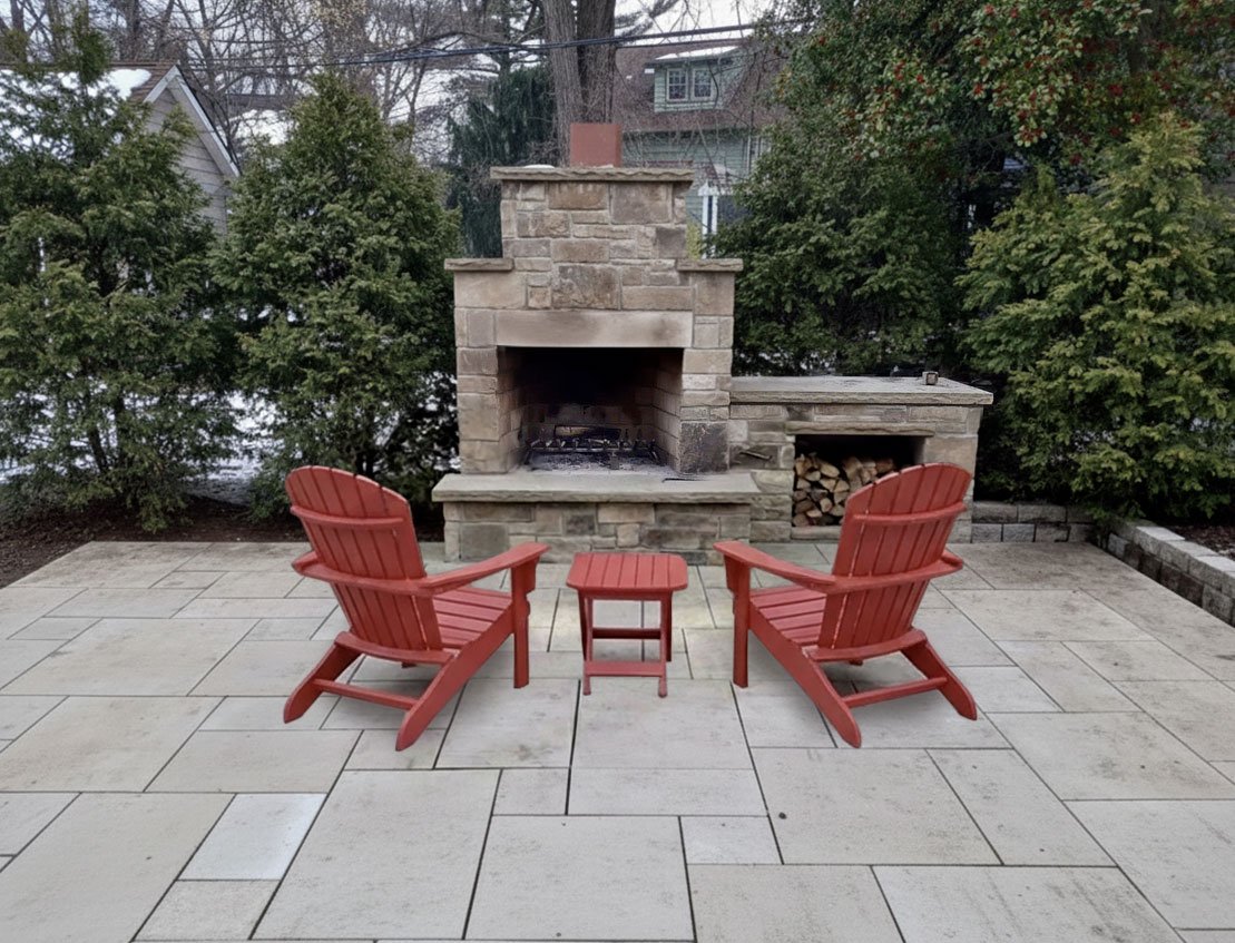 Two red Adirondack chairs with a small matching table between them, set on a tiled patio in front of a stone outdoor fireplace with a wood storage area to the right, surrounded by bushes and trees.