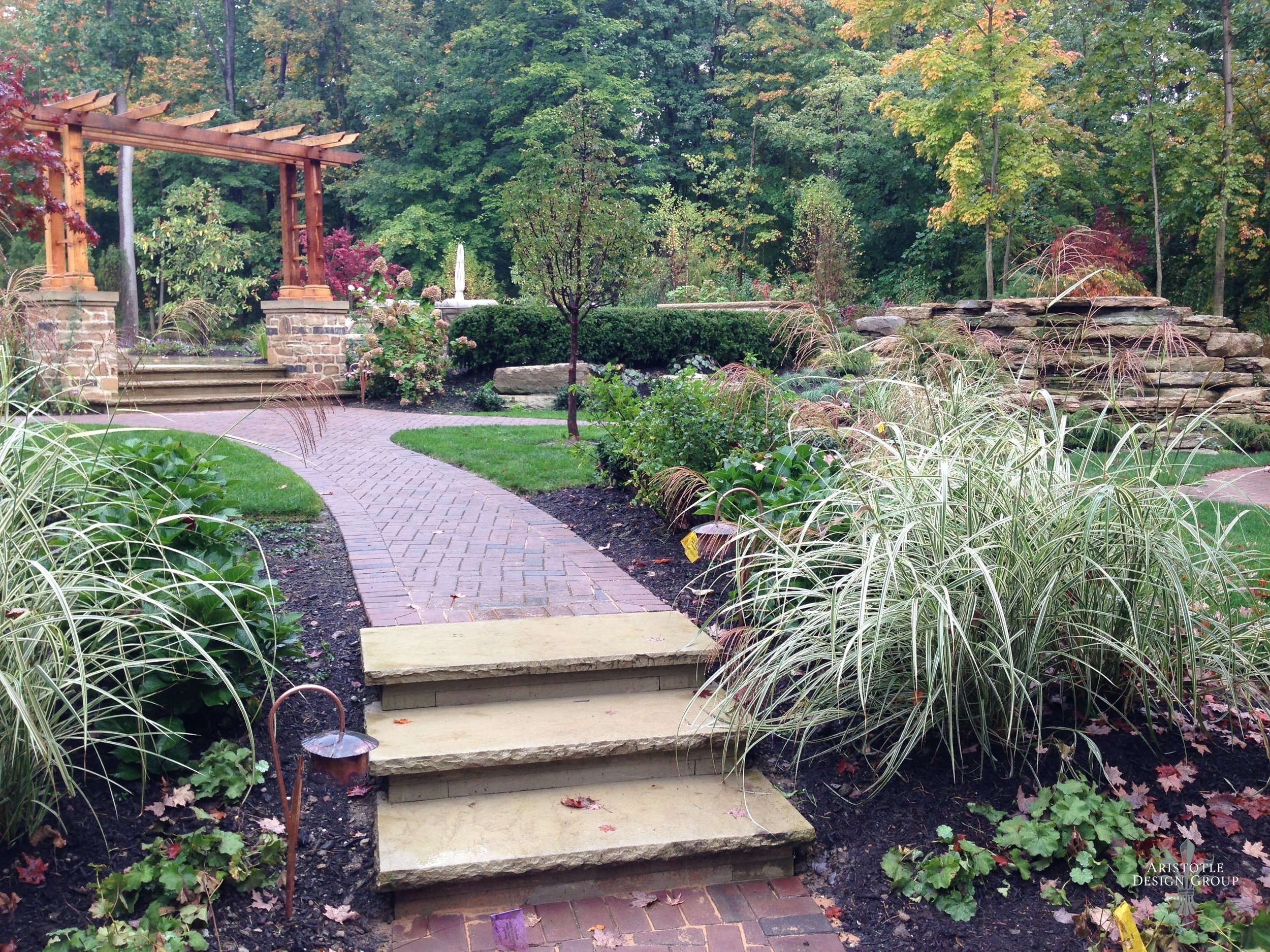 A landscaped garden with a brick pathway, stone steps, and a wooden pergola surrounded by plants, trees, and bushes.