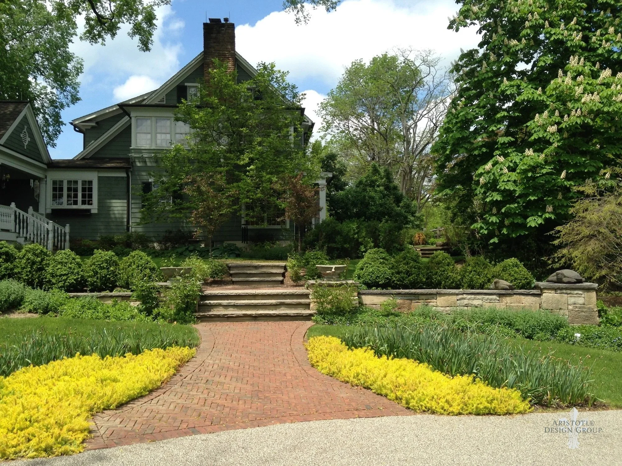 Front yard with brick pathway, yellow and green plants, and a large green house surrounded by trees and bushes under partly cloudy sky.
