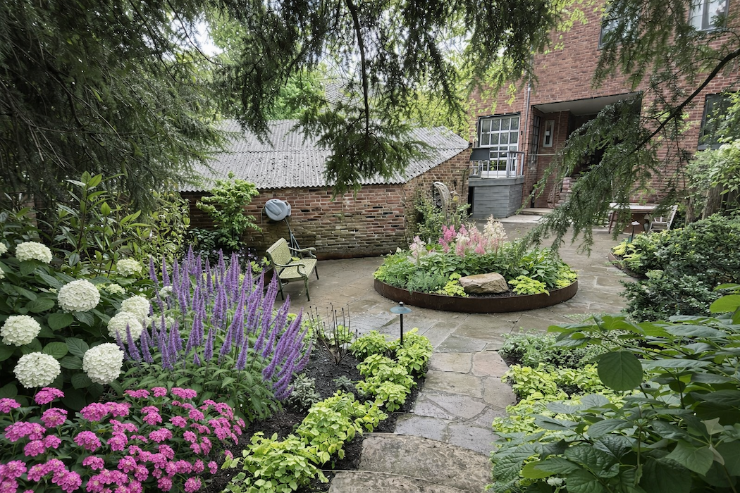 A lush garden courtyard with colorful flowers, a stone pathway, a bench, and a brick house in the background.