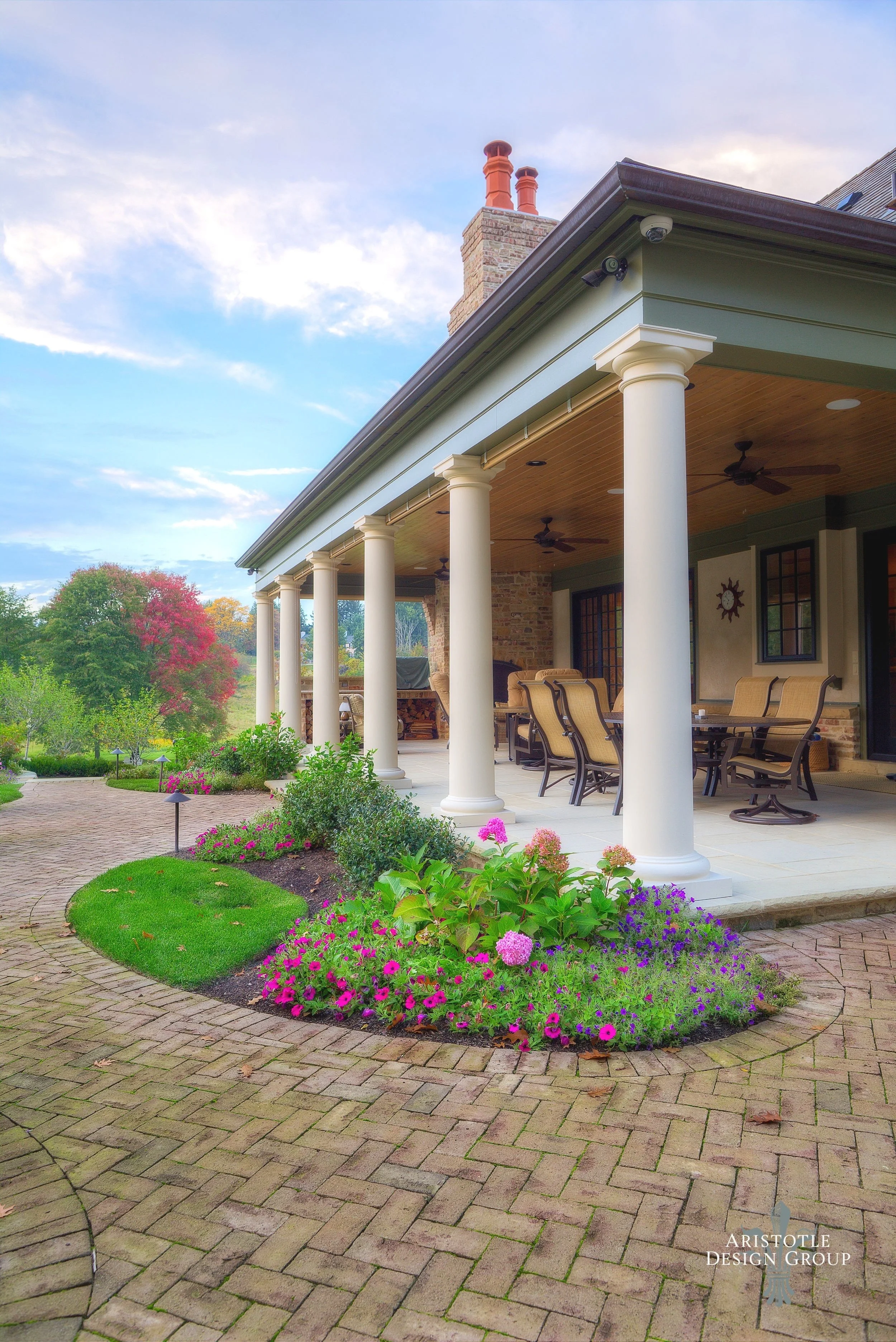 A spacious covered porch of a house with outdoor furniture, surrounded by a landscaped garden with colorful flowers, green bushes, and trees with autumn foliage, under a partly cloudy sky.
