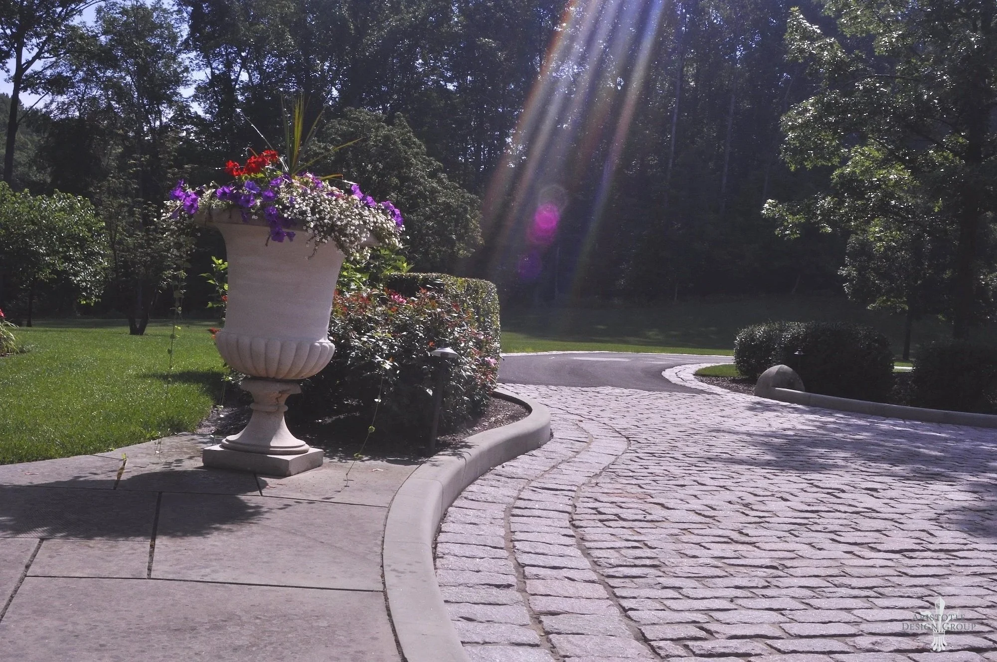A large white ornamented planter with colorful flowers on a sidewalk near a curved cobblestone road, with sunlight streaming through trees in a lush green park.