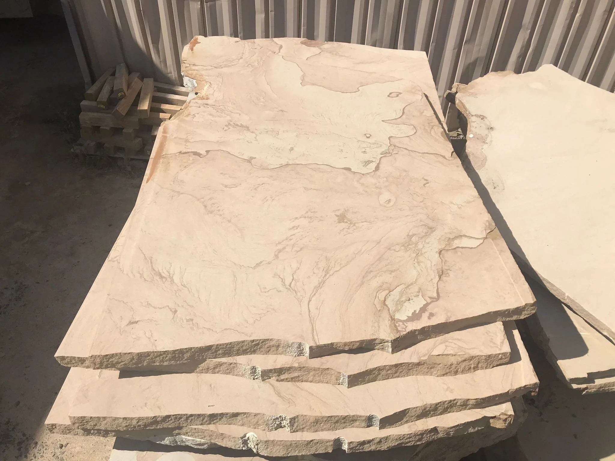 Stack of large, irregularly shaped flat stone slabs with natural beige and light brown marbled patterns, stored outdoors against a corrugated metal wall.