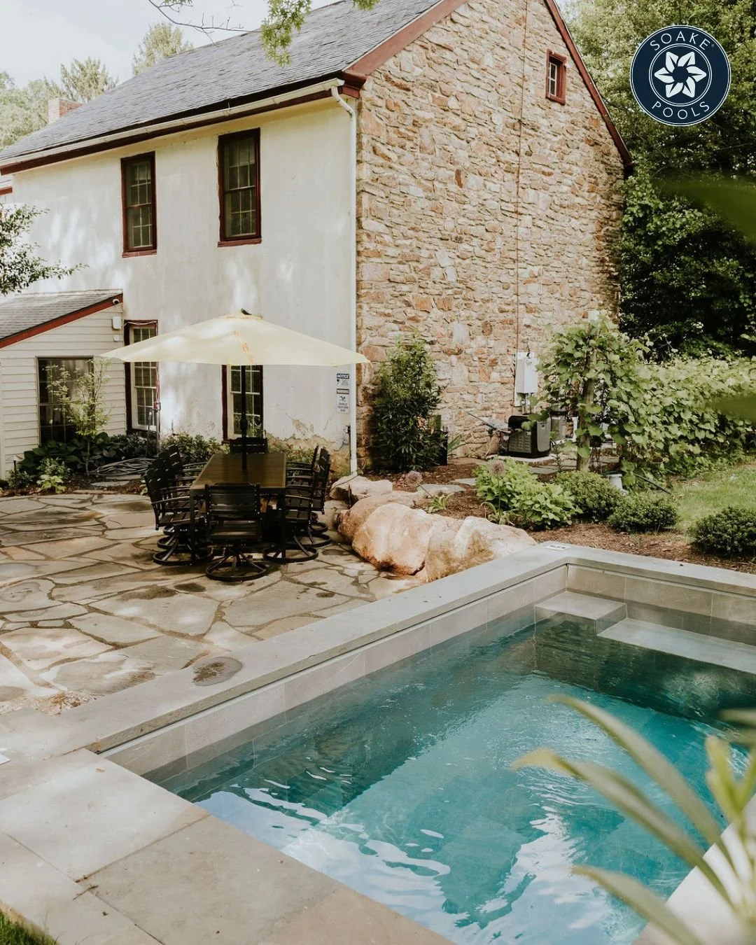 Backyard with swimming pool, patio table with black chairs under a white umbrella, garden plants, and a stone and stucco house with a gable roof.