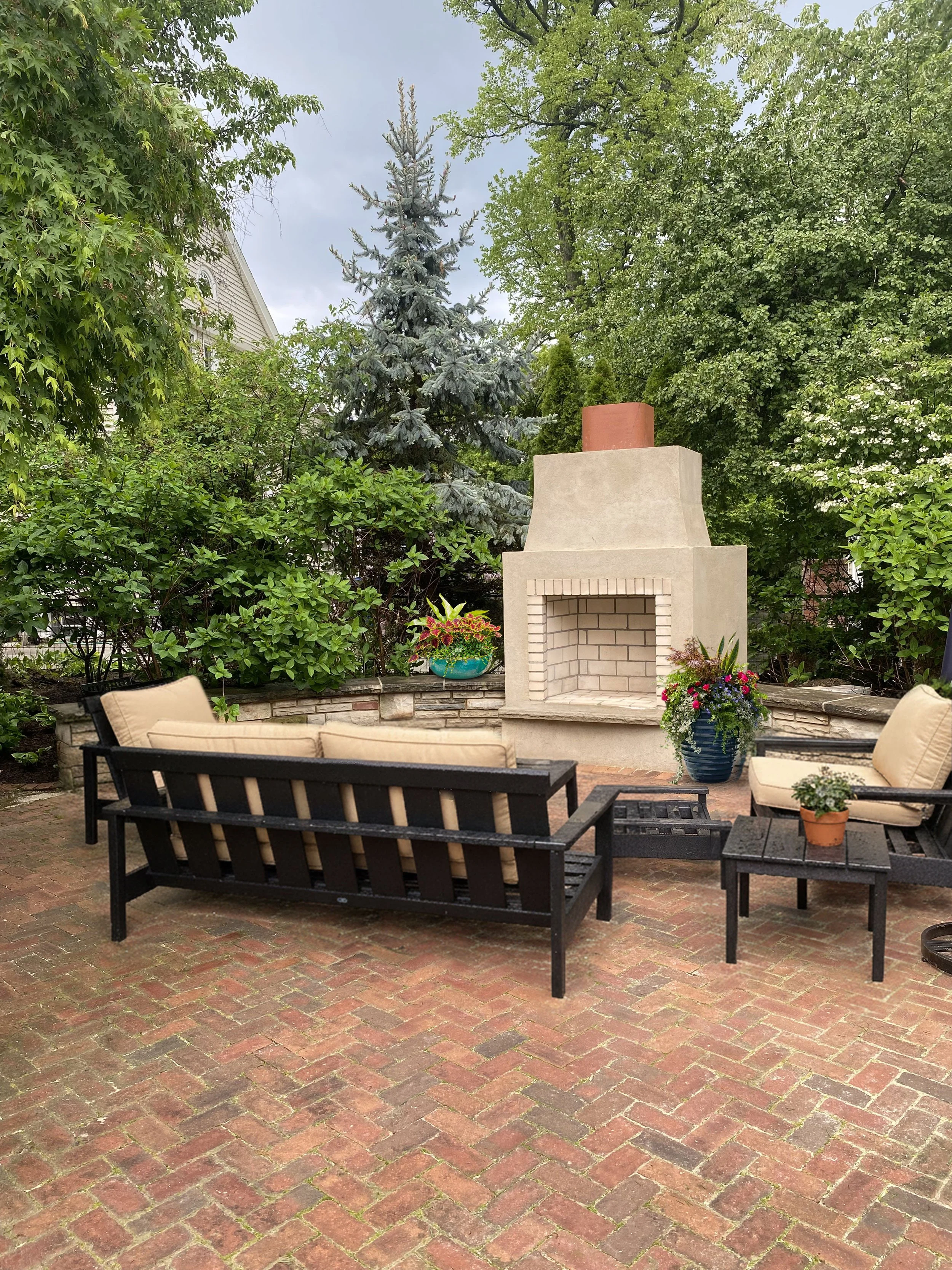 Patio with beige cushioned outdoor furniture, a firepit, and potted plants, surrounded by greenery and trees.