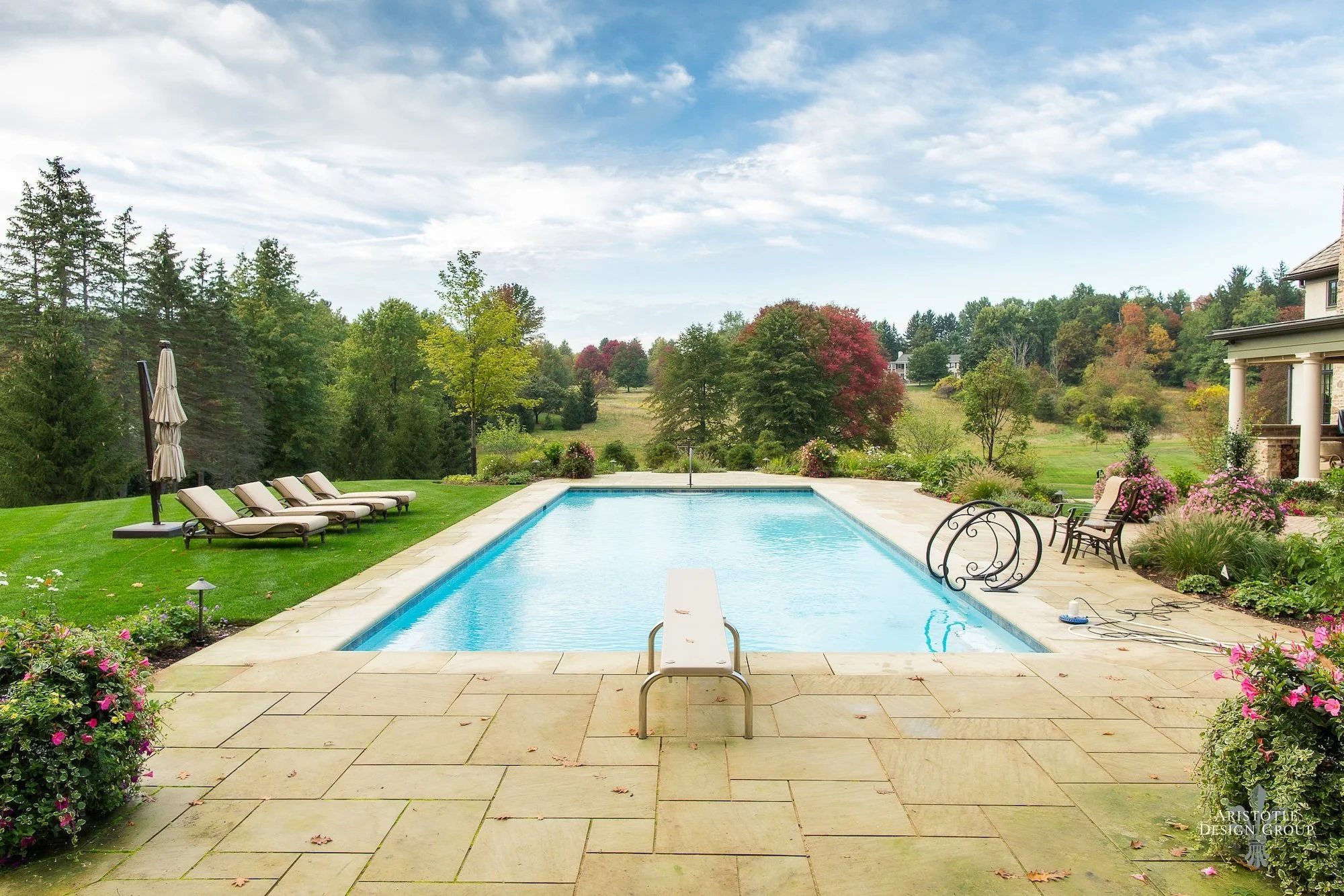 Sunlit backyard with a rectangular swimming pool surrounded by a stone patio, lounge chairs, garden beds with pink flowers, lush green grass, and a scenic landscape with trees and rolling hills under a partly cloudy sky.