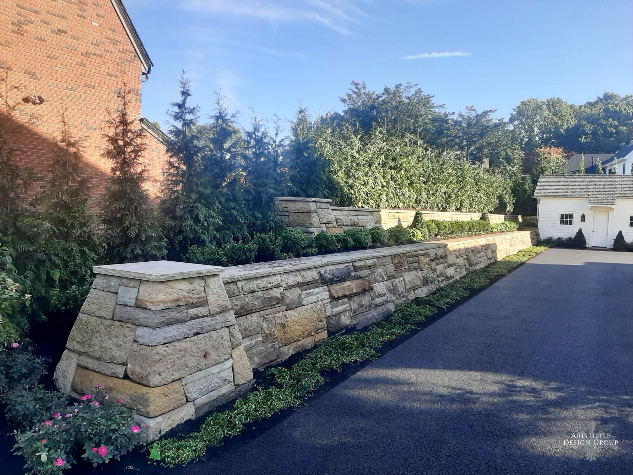 A landscaped backyard with a stone retaining wall, evergreen shrubs, and a tree. There is a brick house on the left and a white shed on the right. The driveway is paved and the sky is blue with some clouds.