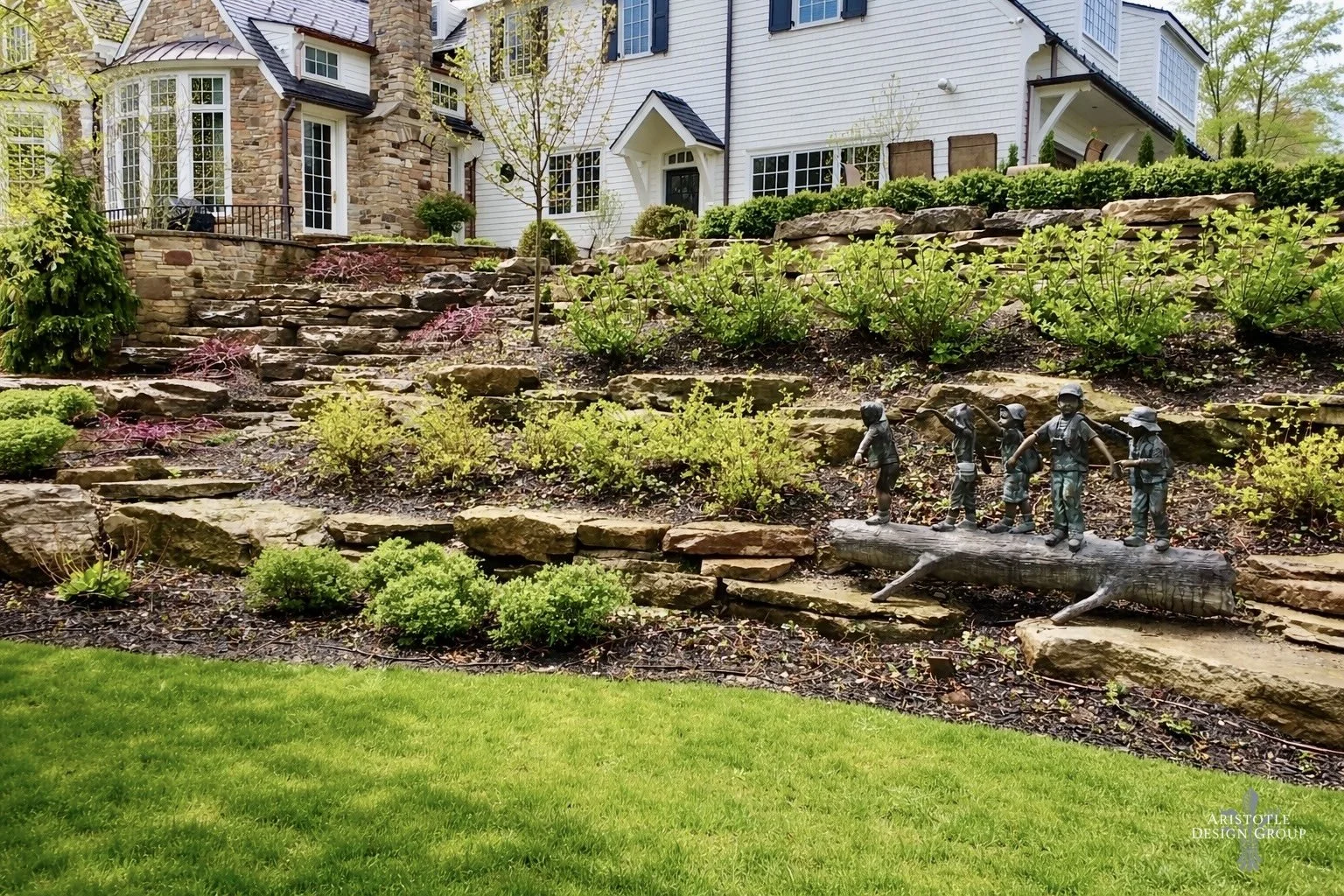 A landscaped garden with layered stone terraces, green shrubs, and small trees in front of a large white house with black shutters. A decorative sculpture of children in firefighter helmets stands on a log in the middle of the garden.