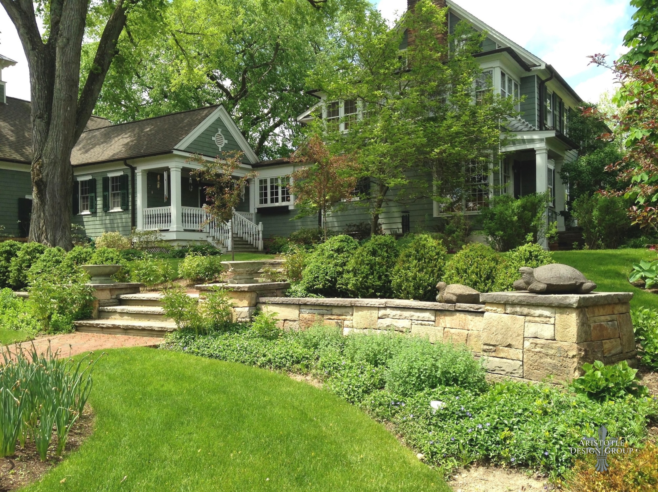 A large, green-colored house with white framing, surrounded by lush landscaping and trees. Stone steps lead up to a porch, and a decorative stone wall with turtle sculptures is in the foreground.