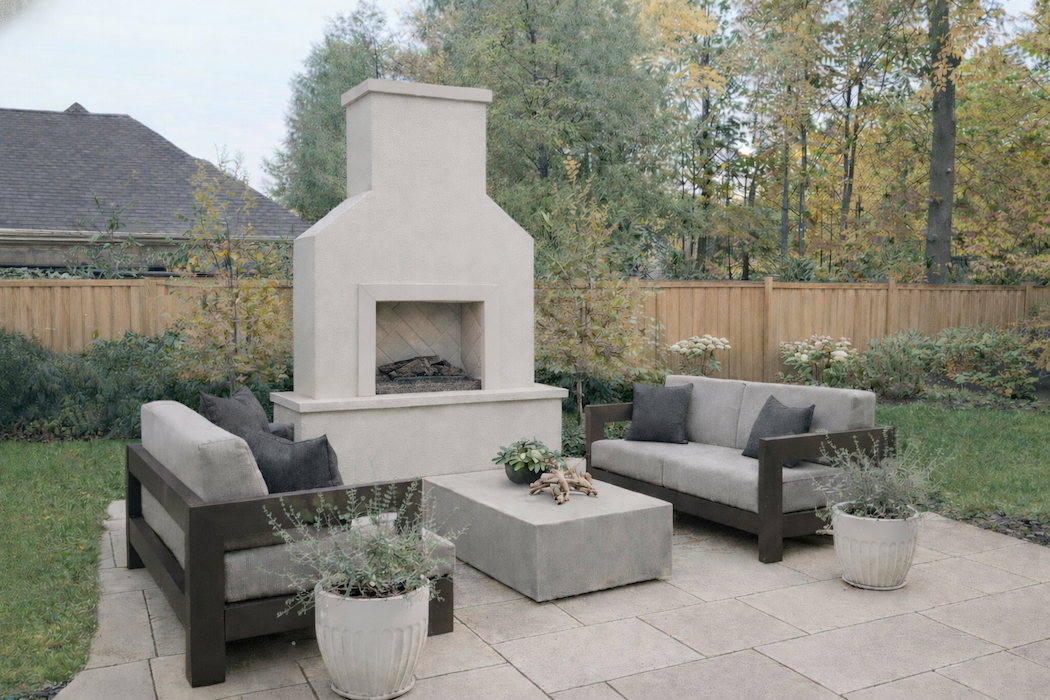 Outdoor patio with a built-in fireplace, two cushioned sofas with dark wooden arms, two gray pillows on each, a large cushioned coffee table, two potted plants, and a wooden fence with trees in the background