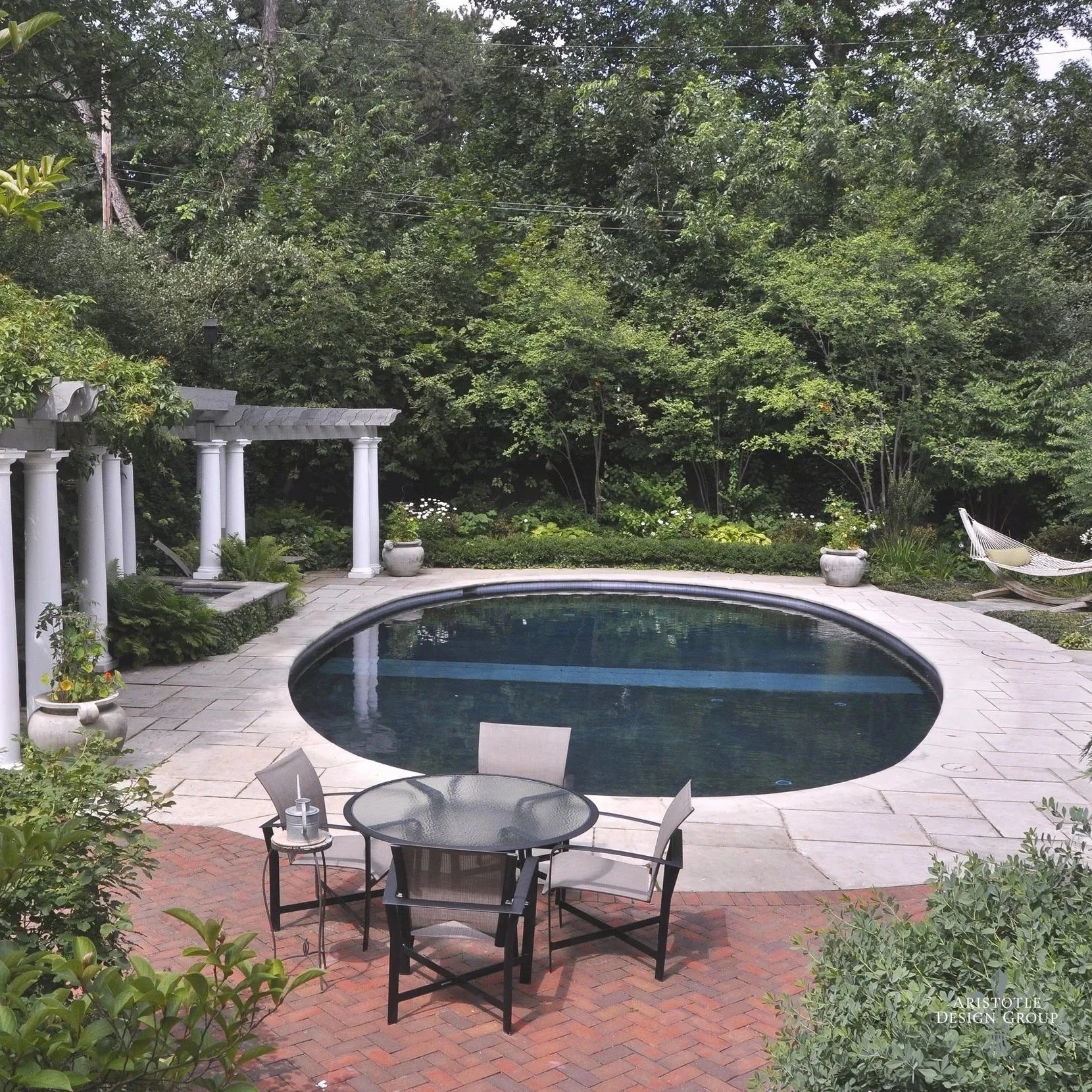 A backyard featuring a small, oval-shaped swimming pool surrounded by a stone patio. There is a black glass outdoor table with four chairs on a brick patio area. Behind the pool, there are lush green trees, potted plants, and a hammock in the corner.