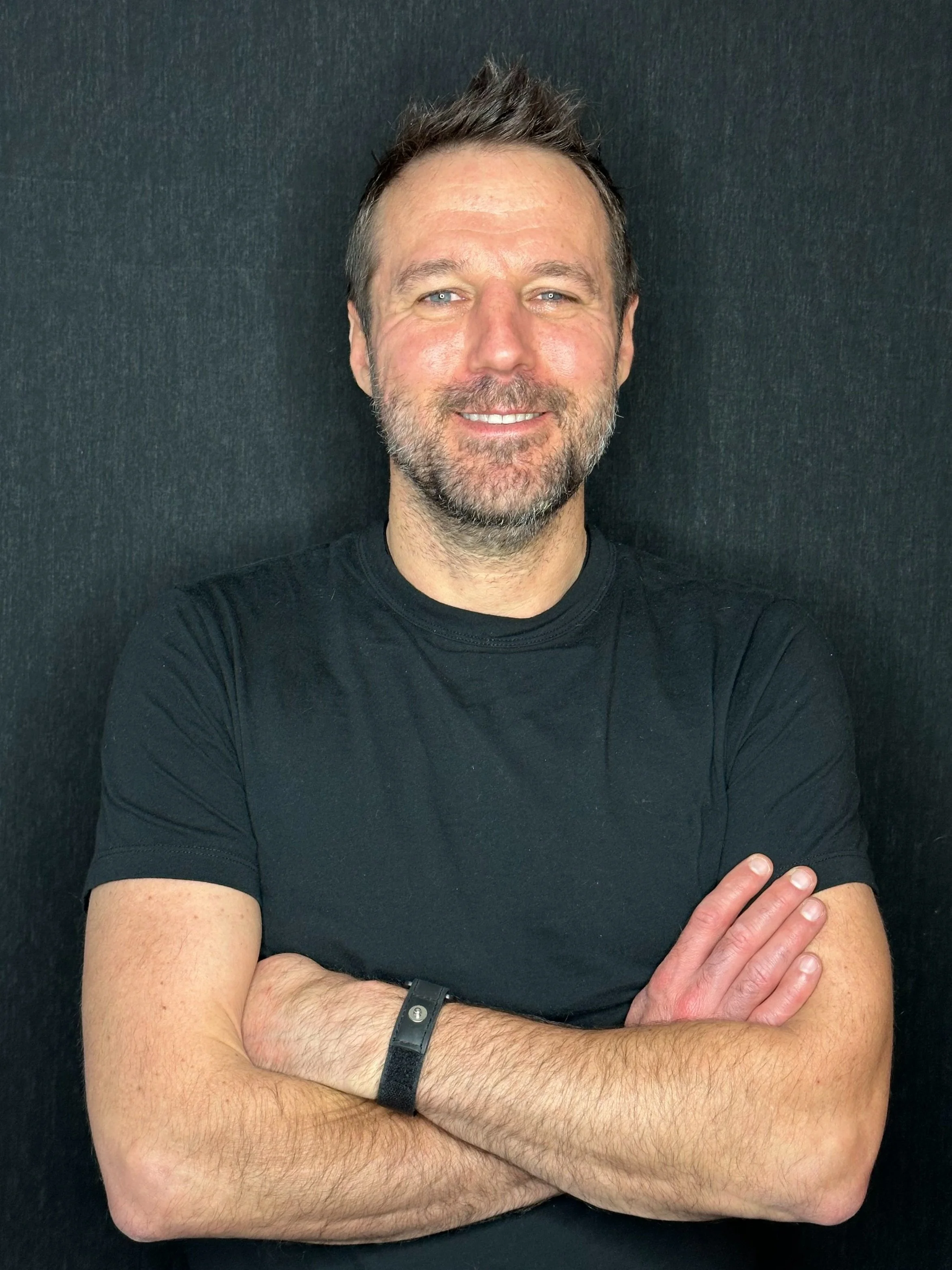 A middle-aged man with short dark hair and a beard, smiling, wearing a black t-shirt, crossing his arms, standing against a dark background.