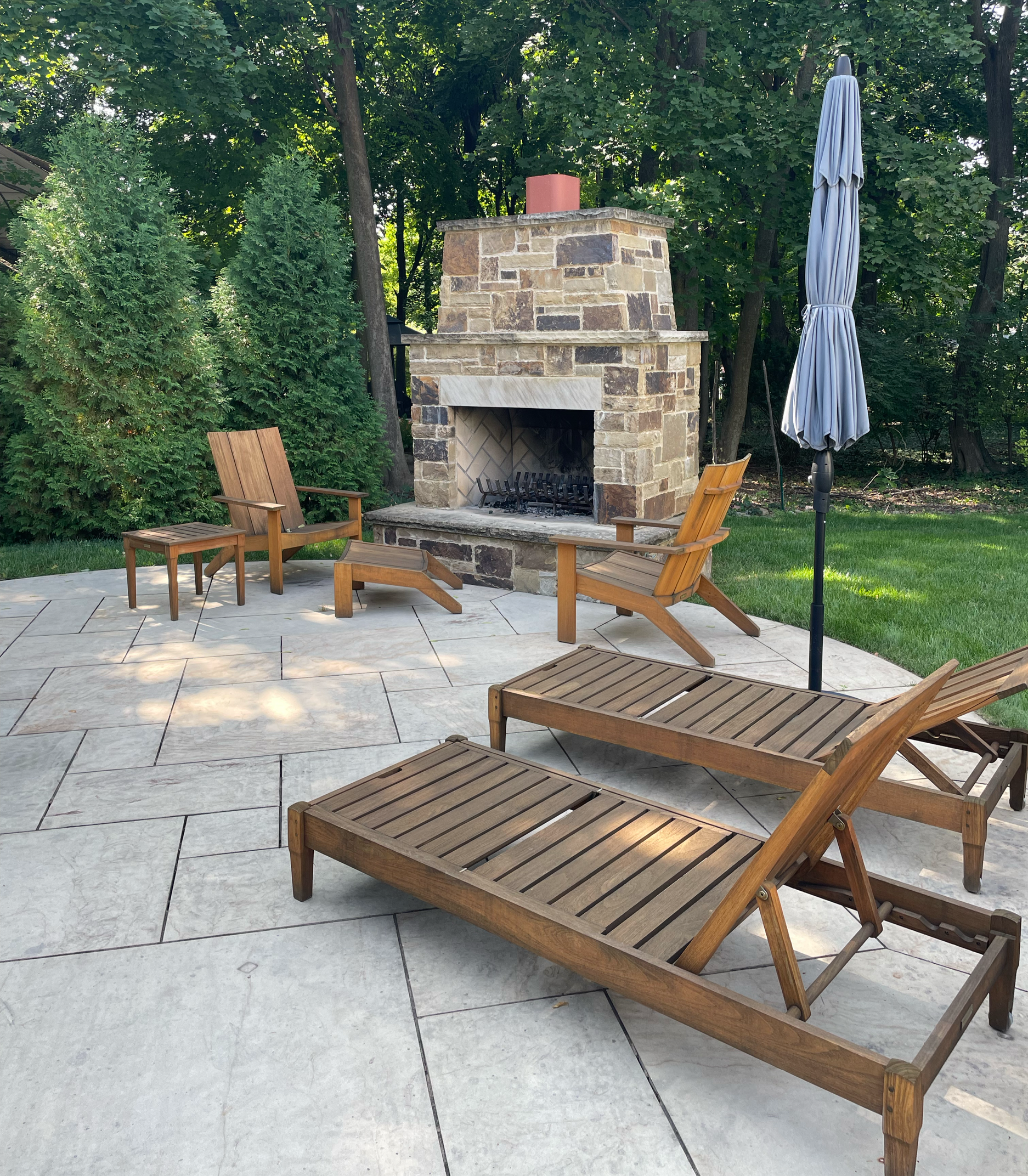 Backyard patio with two wooden lounge chairs, one with a small table, near a stone fireplace, and a closed patio umbrella, with a grassy area and trees in the background.