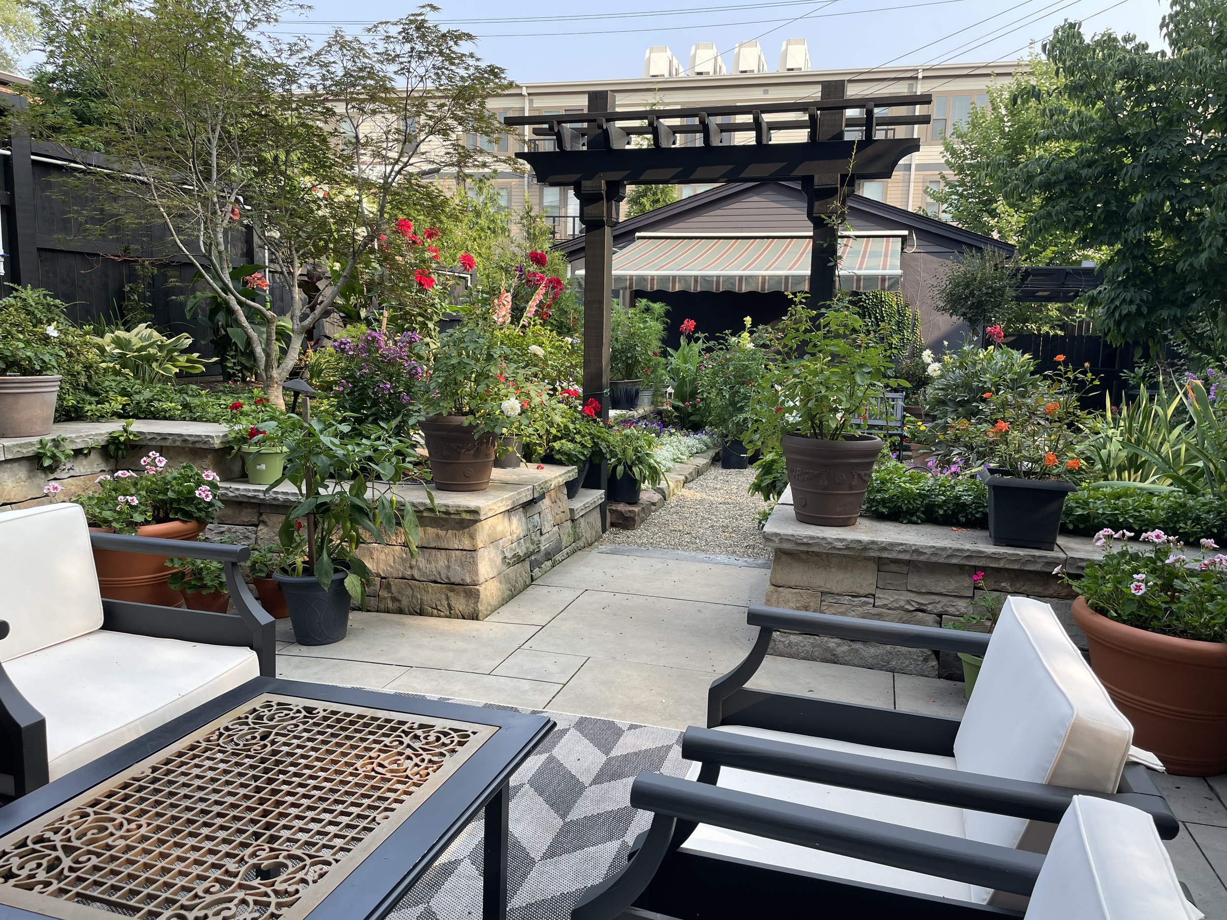 A patio garden with potted flowers and plants, stone retaining walls, seating area with black and white chairs, and a pergola in the background.