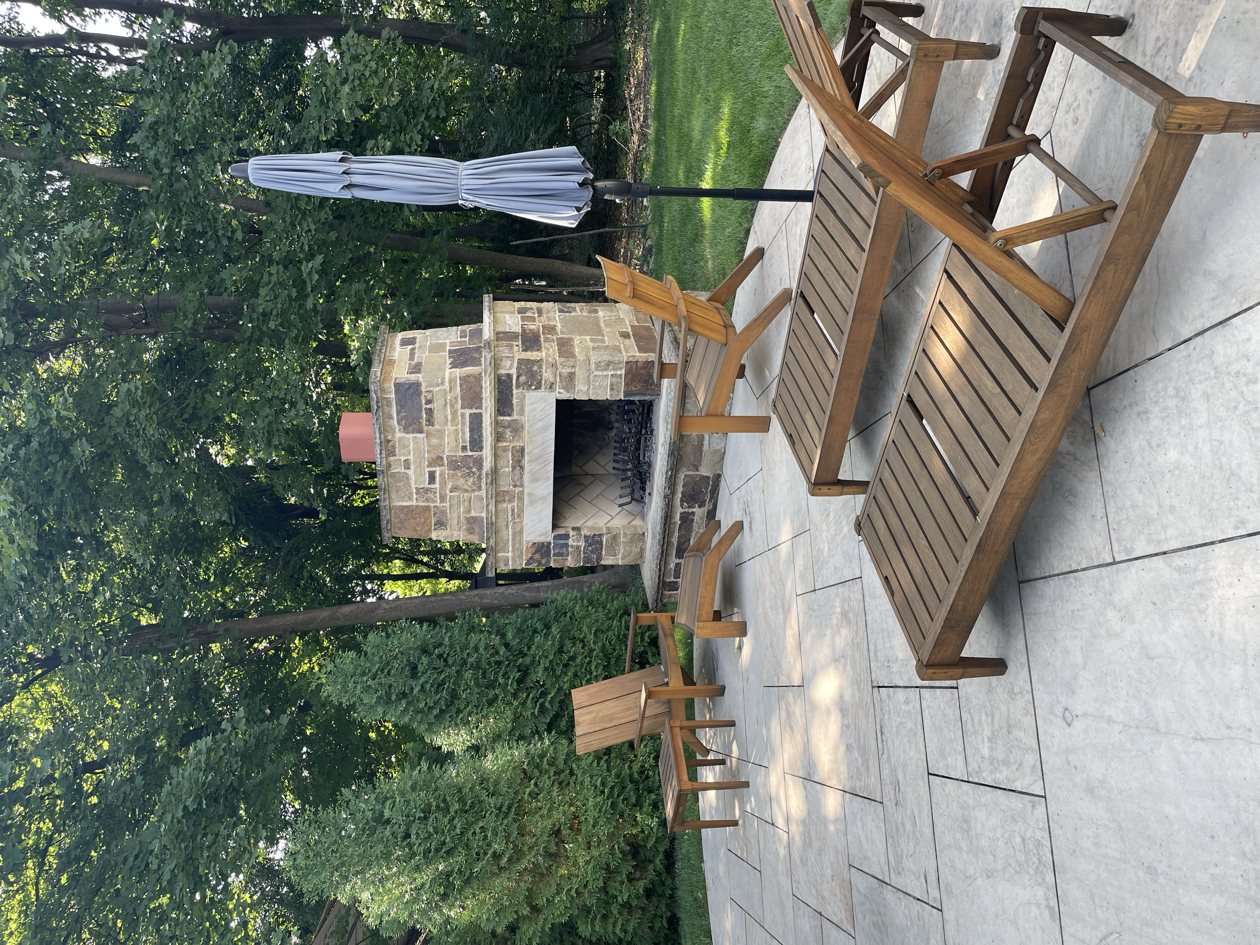 Outdoor patio area with wooden furniture, including a glass-top table, chairs, and a sun umbrella, in front of a stone fireplace, surrounded by green trees and grass.