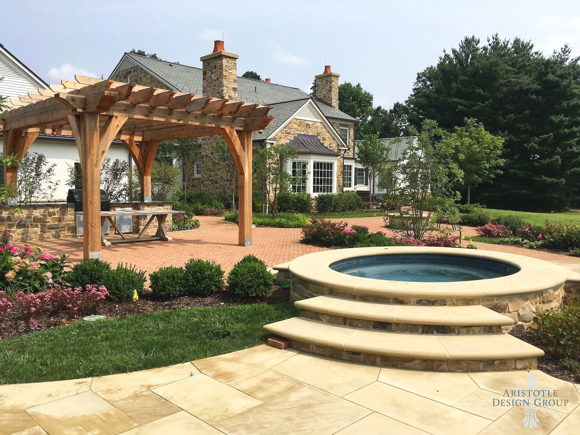 Backyard with a stone house, a pergola, a hot tub with steps, and landscaped garden with flowers and trees.