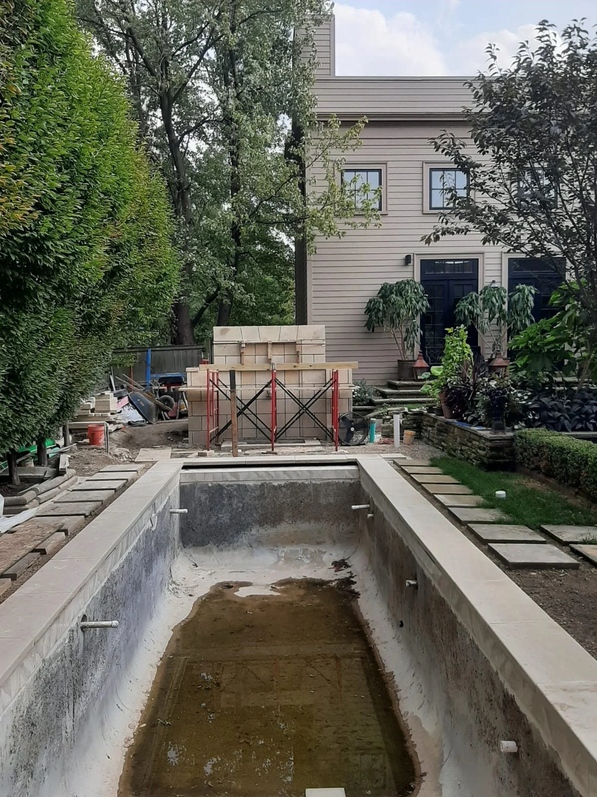 An empty swimming pool under construction in a backyard, with construction materials and scaffolding, surrounded by greenery and a beige house with black doors and windows.