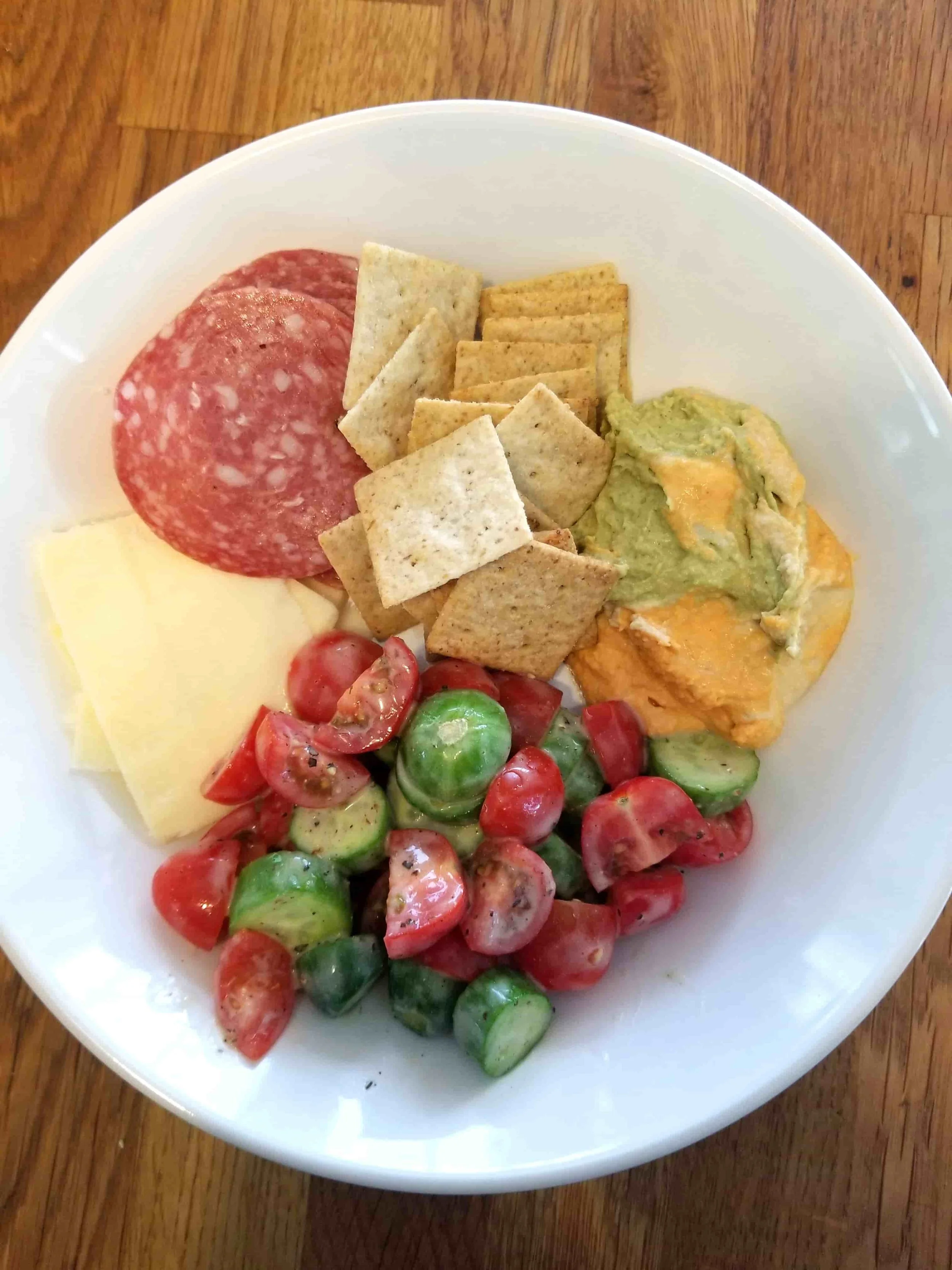 A meal of snacks including cheese, salami, hummus, crackers, and a cucumber and tomato salad.
