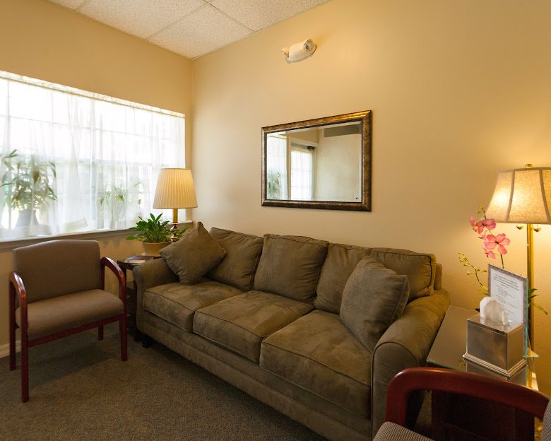 Waiting room with light colored walls, a large window with white curtains, a beige sofa with pillows, a side table with a lamp, a mirror on the wall, and a potted plant.