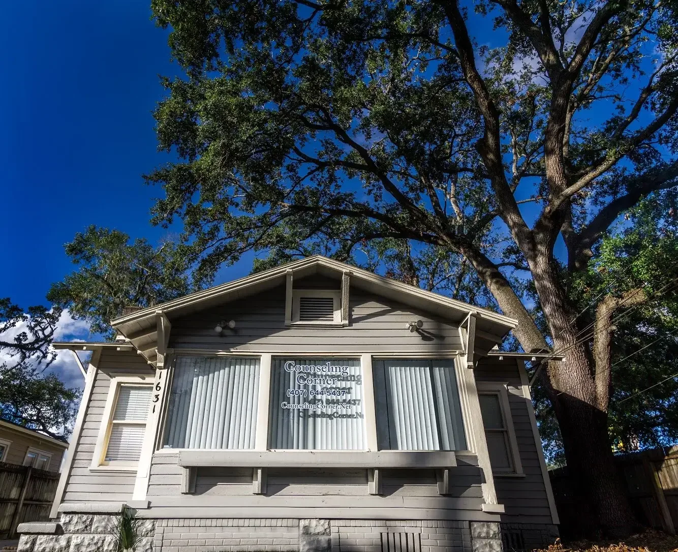 Counseling Corner Orlando location, a small historic house with a large tree behind it, advertising counseling services on its front window, located at 1631 Hillcrest St. 32803, with contact number 407-644-5437 under a blue sky.