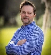 A man with short brown hair, wearing a blue checkered shirt, standing outdoors with trees and grass in the background.