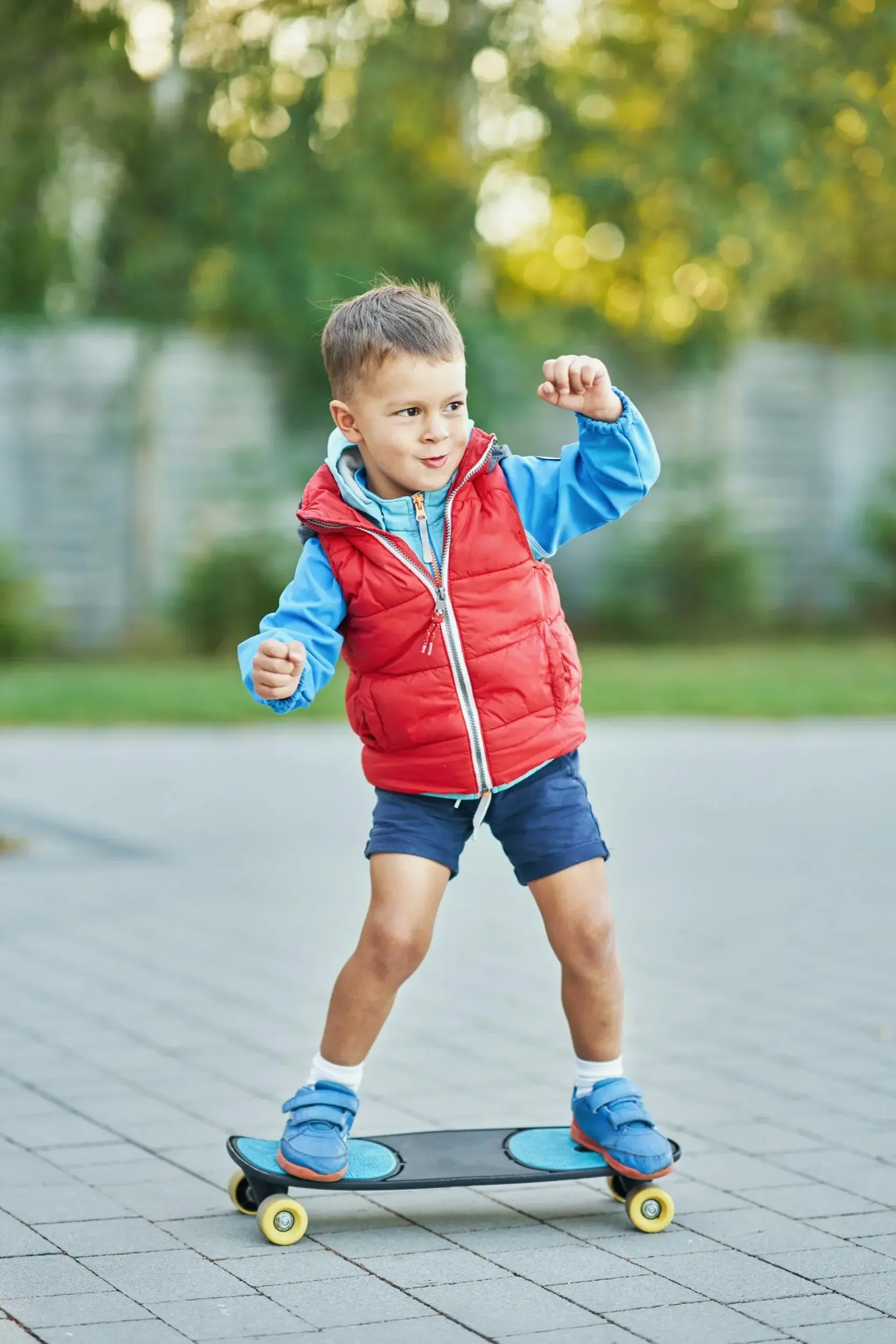 A young boy wearing a red vest, blue jacket, and shorts is standing on a skateboard on paved ground outdoors. He is flexing his right arm and smiling, surrounded by a blurred background of trees and sunlight.