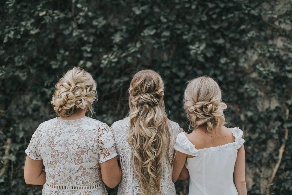 Three blonde woman in wedding wear facing away from the camera, showing their elaborately braided hair