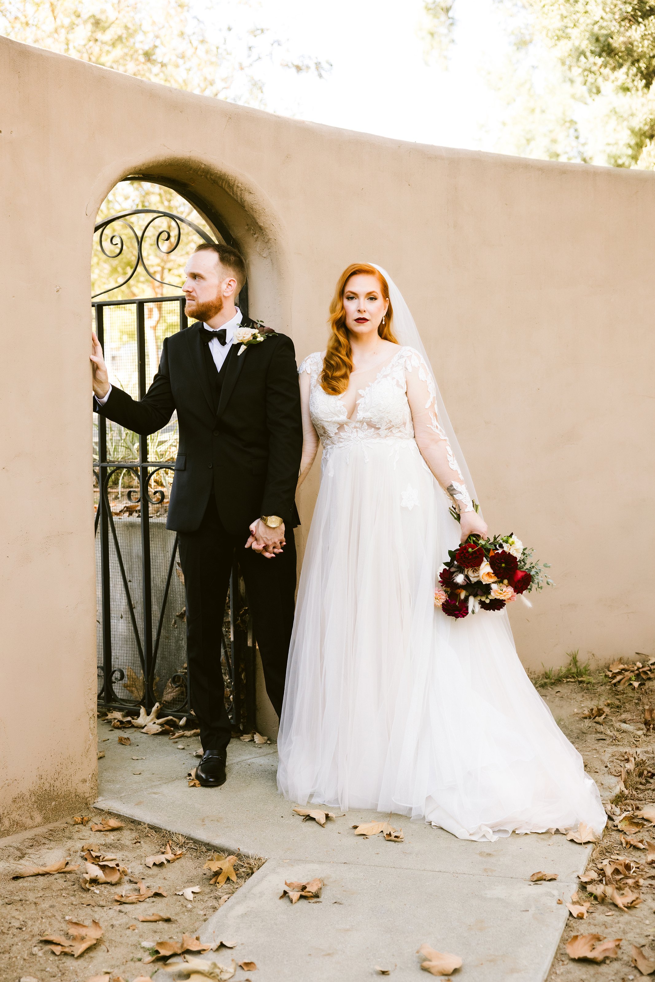 Bride and groom holding hands for a wedding photo outside, with the bride in a white gown holding a bouquet and the groom in a black suit and tie, standing near a beige wall with an arched opening.