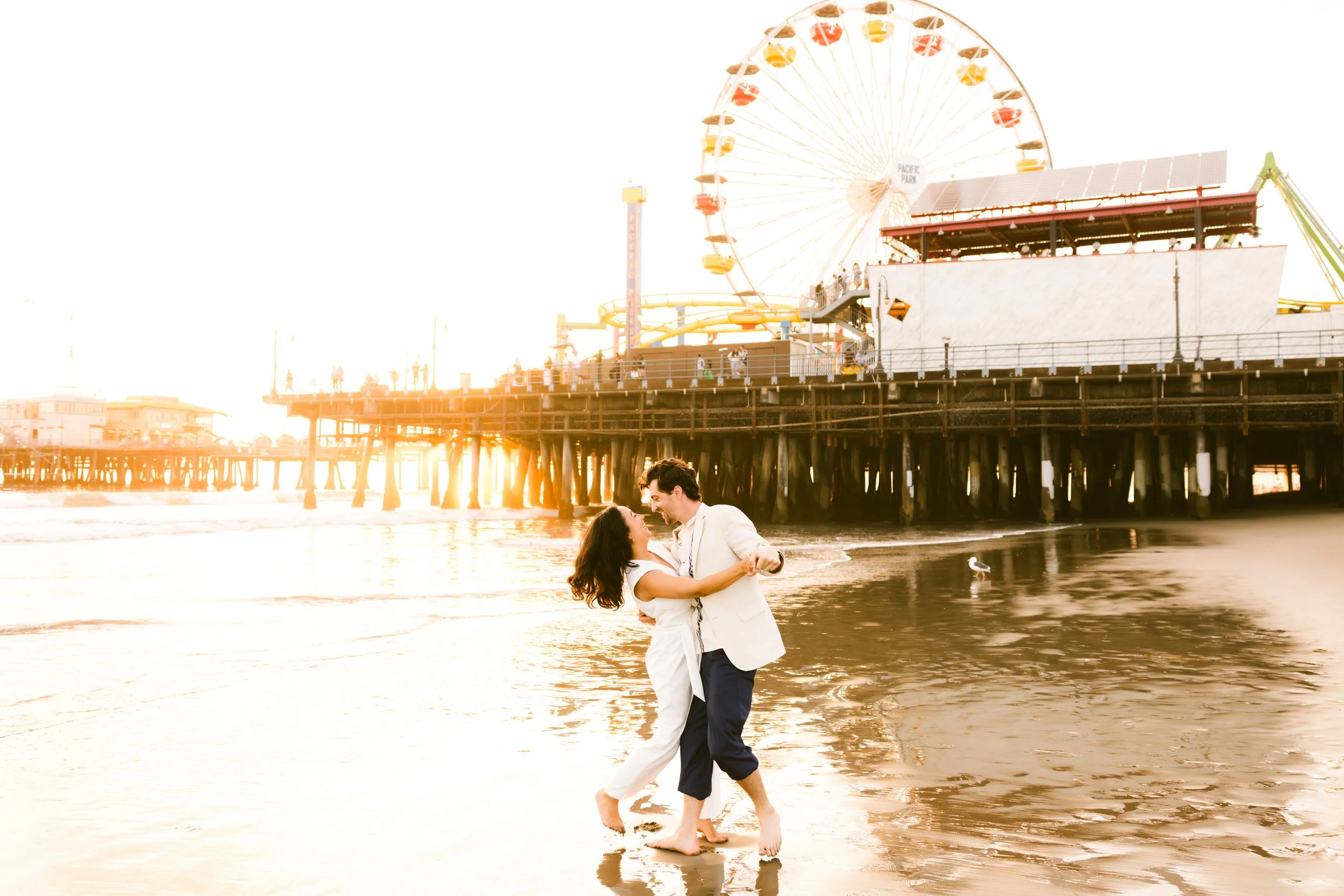 A couple dancing on the beach at sunset near a pier with a Ferris wheel in the background.