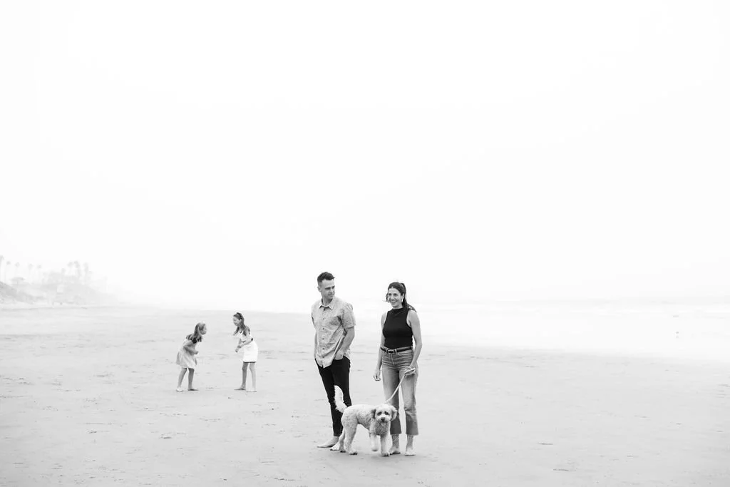 A couple walking their dog on the beach, with two children playing in the background.