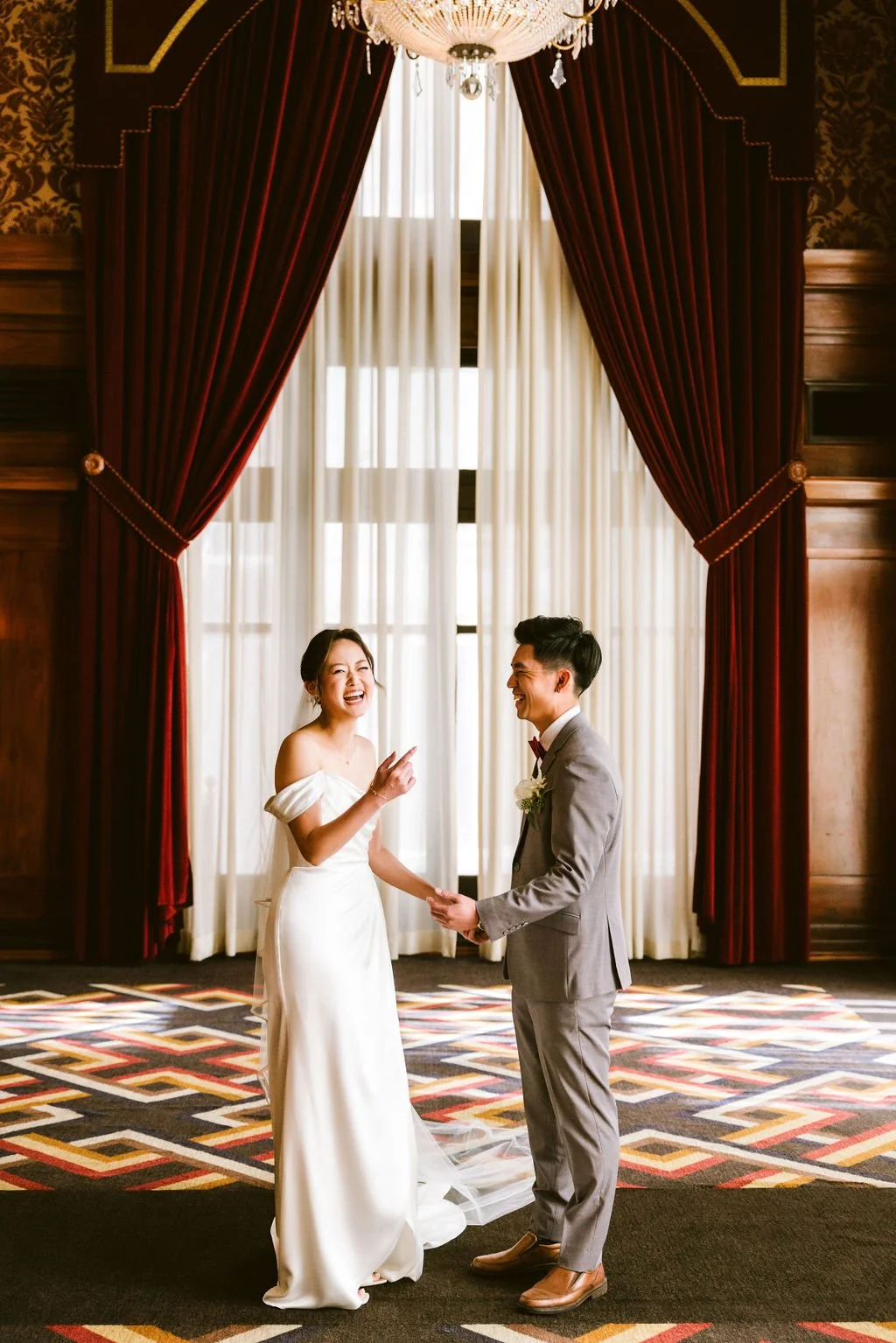 A bride and groom holding hands, smiling, with the bride pointing upwards, in a luxurious room with red curtains and a chandelier, after their wedding ceremony.