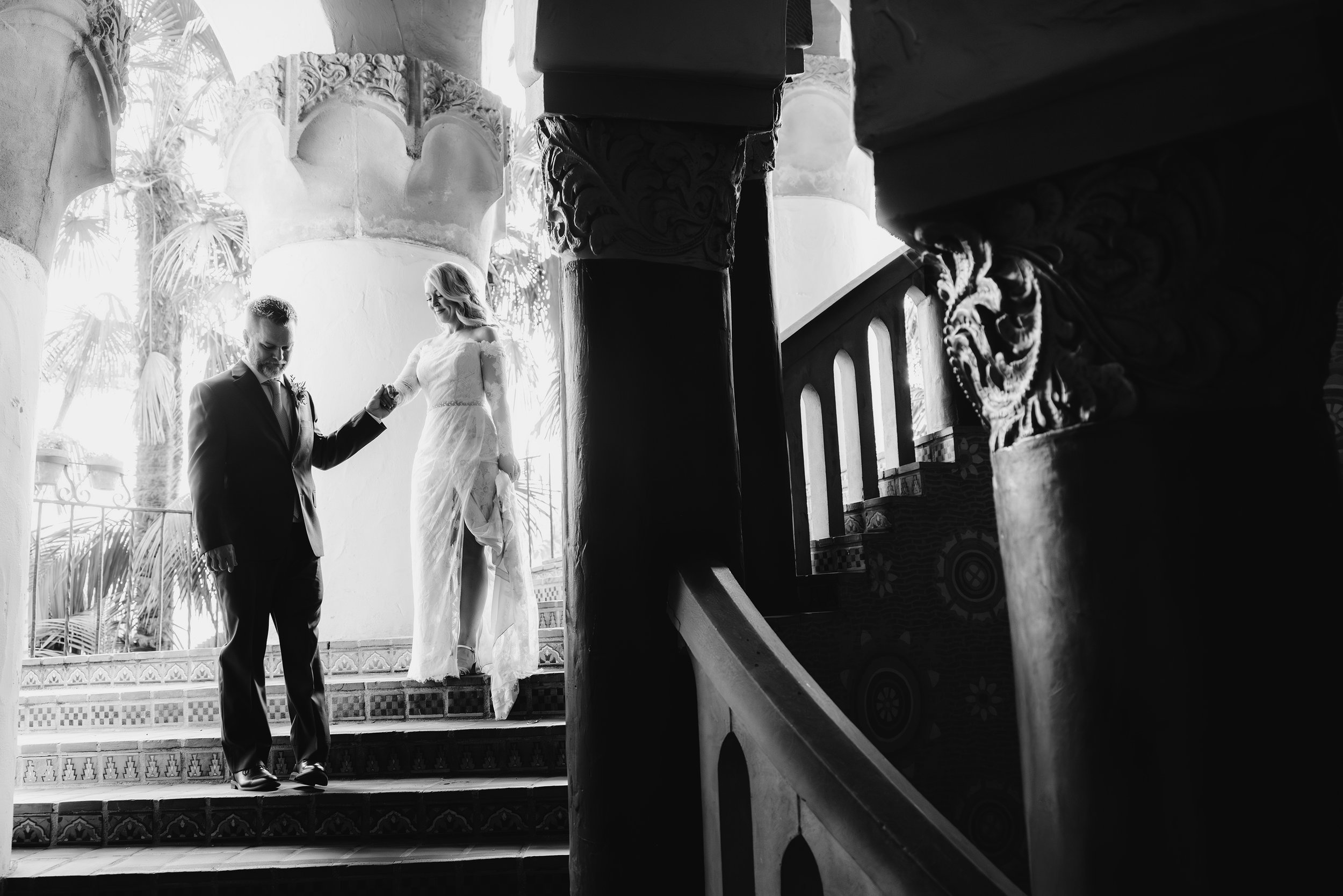 A black and white photo capturing a man and a woman on a decorated staircase inside a building. The woman, dressed in a lace wedding gown, is holding the man's hand and looking at him. The man is dressed in a suit and is looking downward.