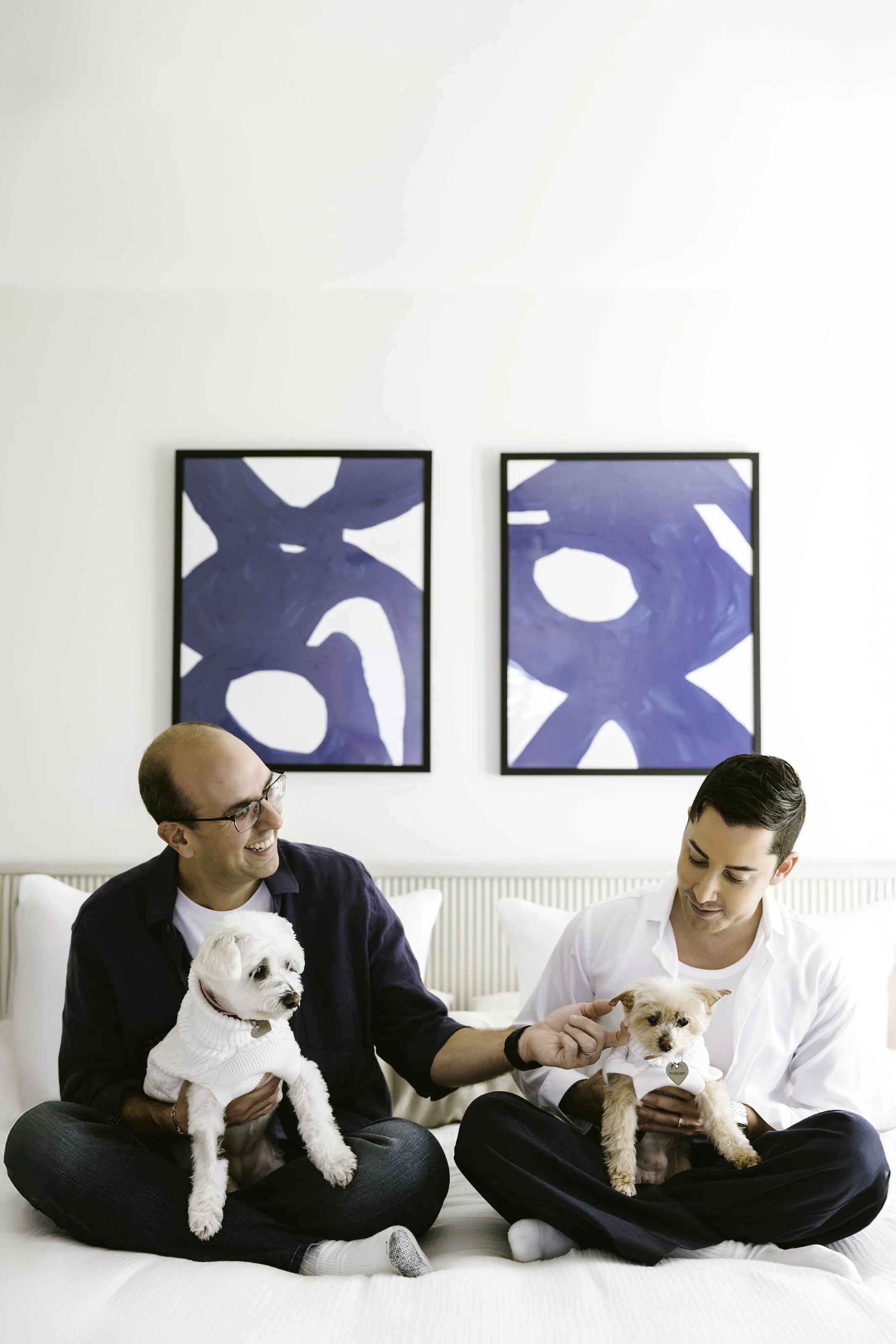 Two men sitting on a bed, each holding a small dog, with two abstract paintings hanging on a white wall behind them.