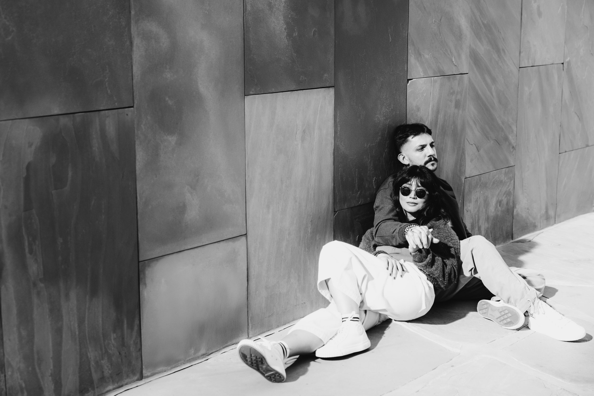 Two young adults sitting on the ground against a tiled wall. The person in front is wearing sunglasses, a dark sweater, and light pants, while the person behind has a dark shirt and mustache. Both appear relaxed, with the person in front holding onto the arm of the person behind.