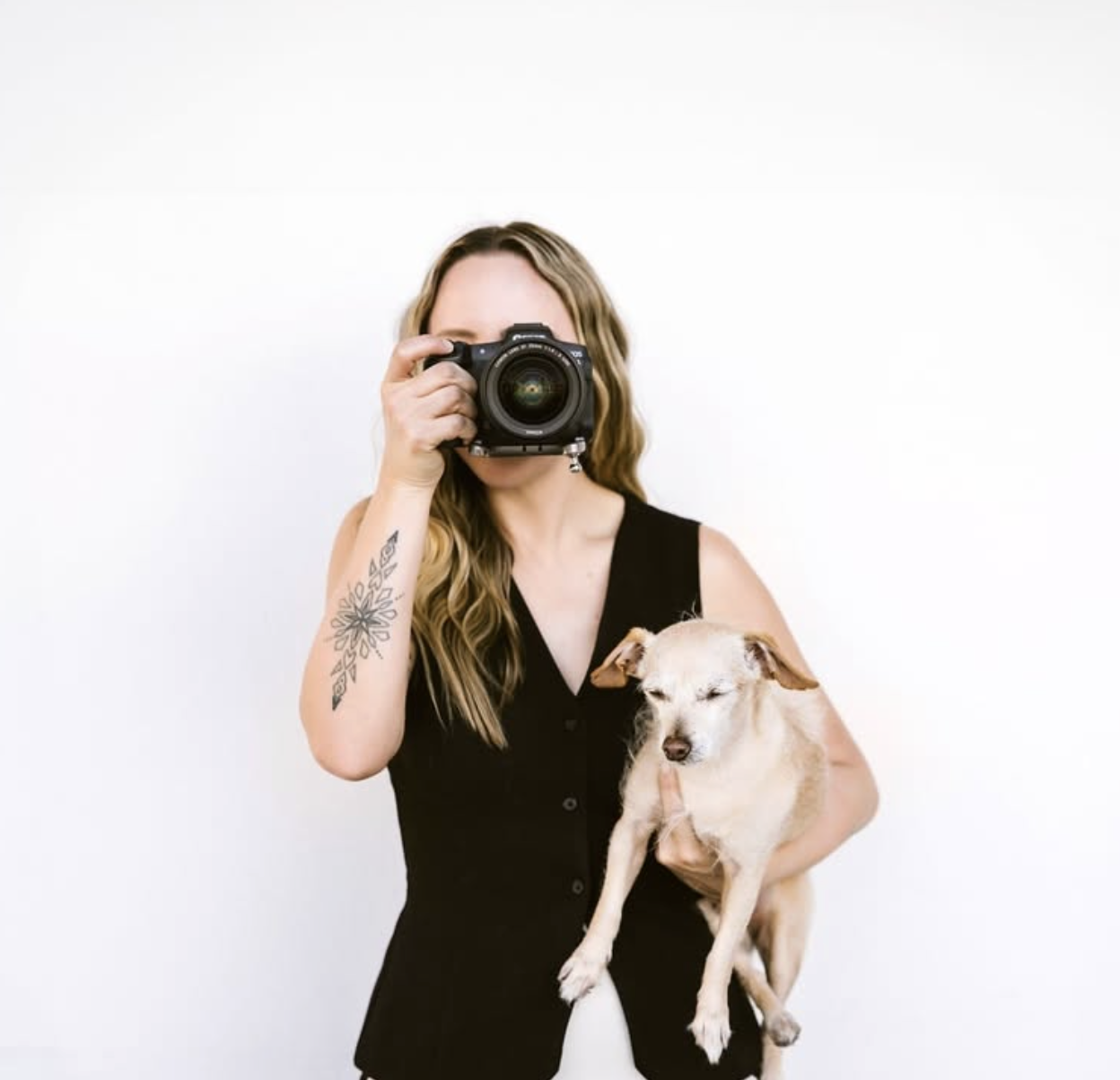 Woman with long wavy hair, tattoo on her left arm, holding a small dog, taking a photo with a camera in front of a plain white background.
