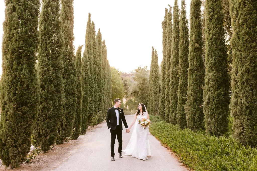 A bride and groom holding hands and walking along a tree-lined path during their wedding day. The bride wears a white wedding dress and holds a bouquet, while the groom is dressed in a black tuxedo with a bow tie.