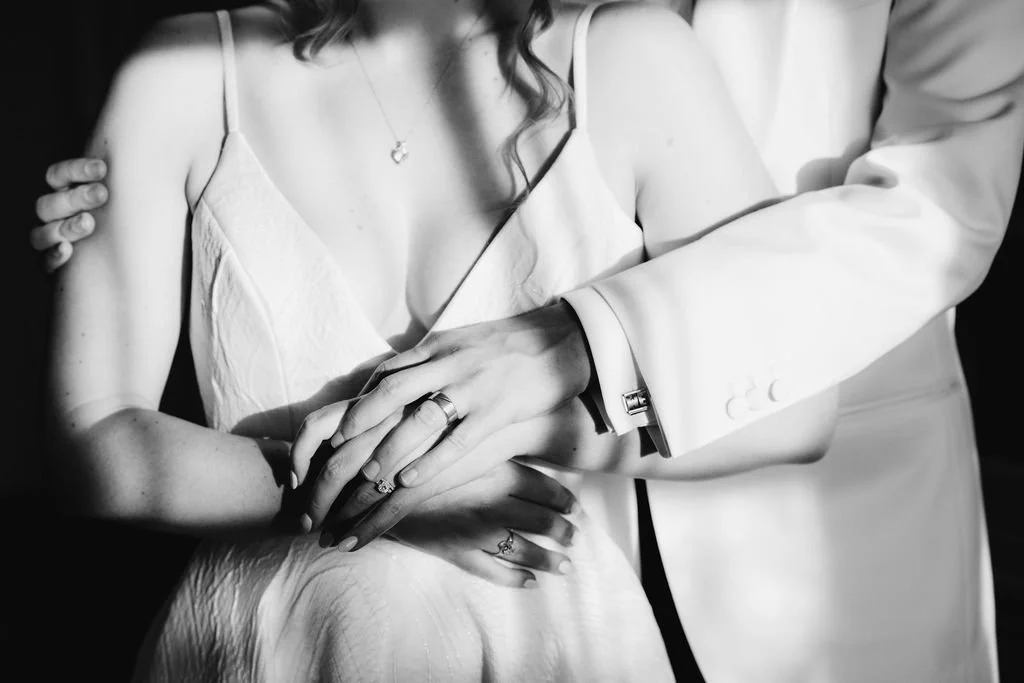 A close-up black and white photo of a bride and groom holding hands, showing wedding rings, with the bride wearing a satin dress and the groom in a suit.
