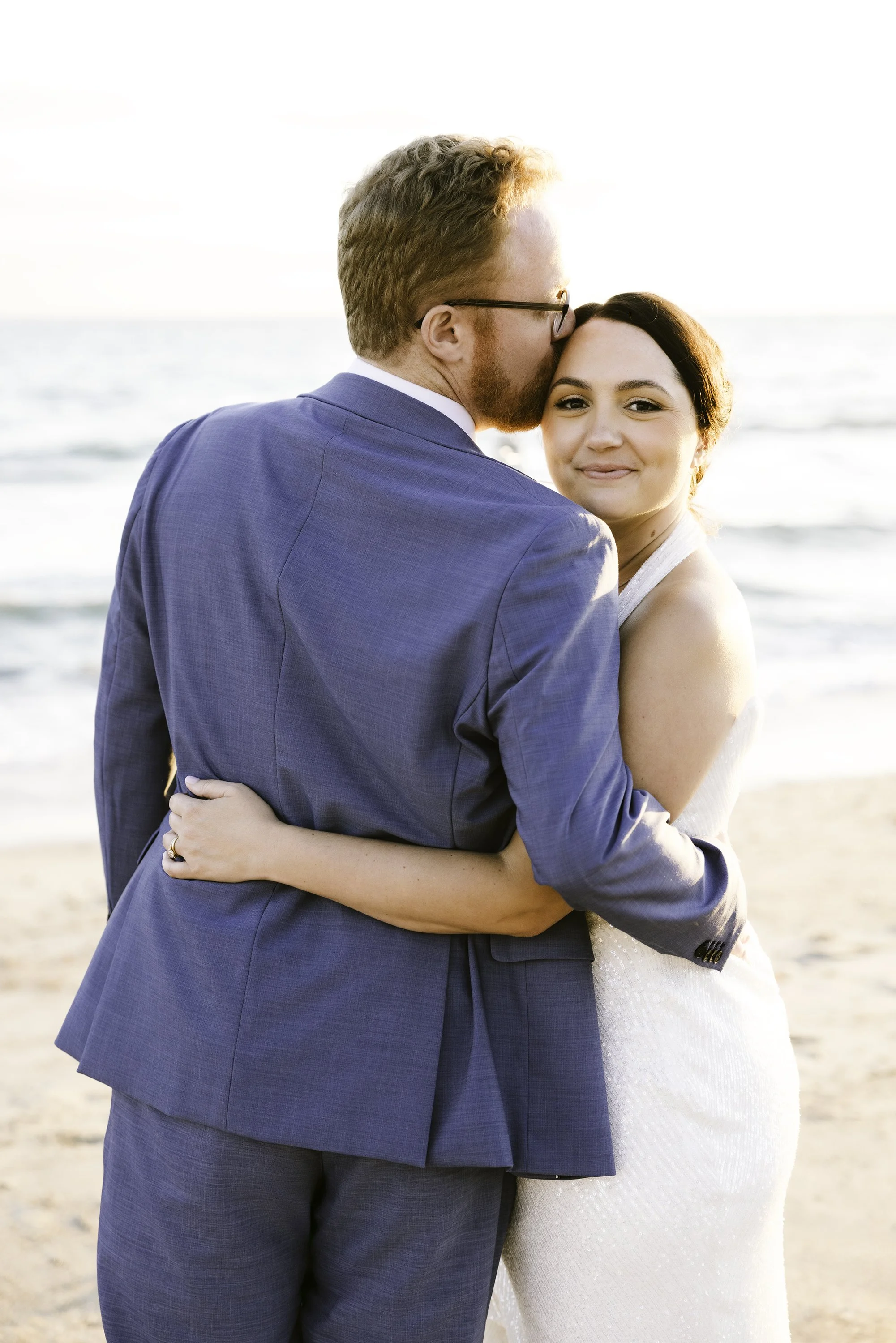 A couple embracing on the beach at sunset, the man kissing the woman on her temple, both smiling softly.