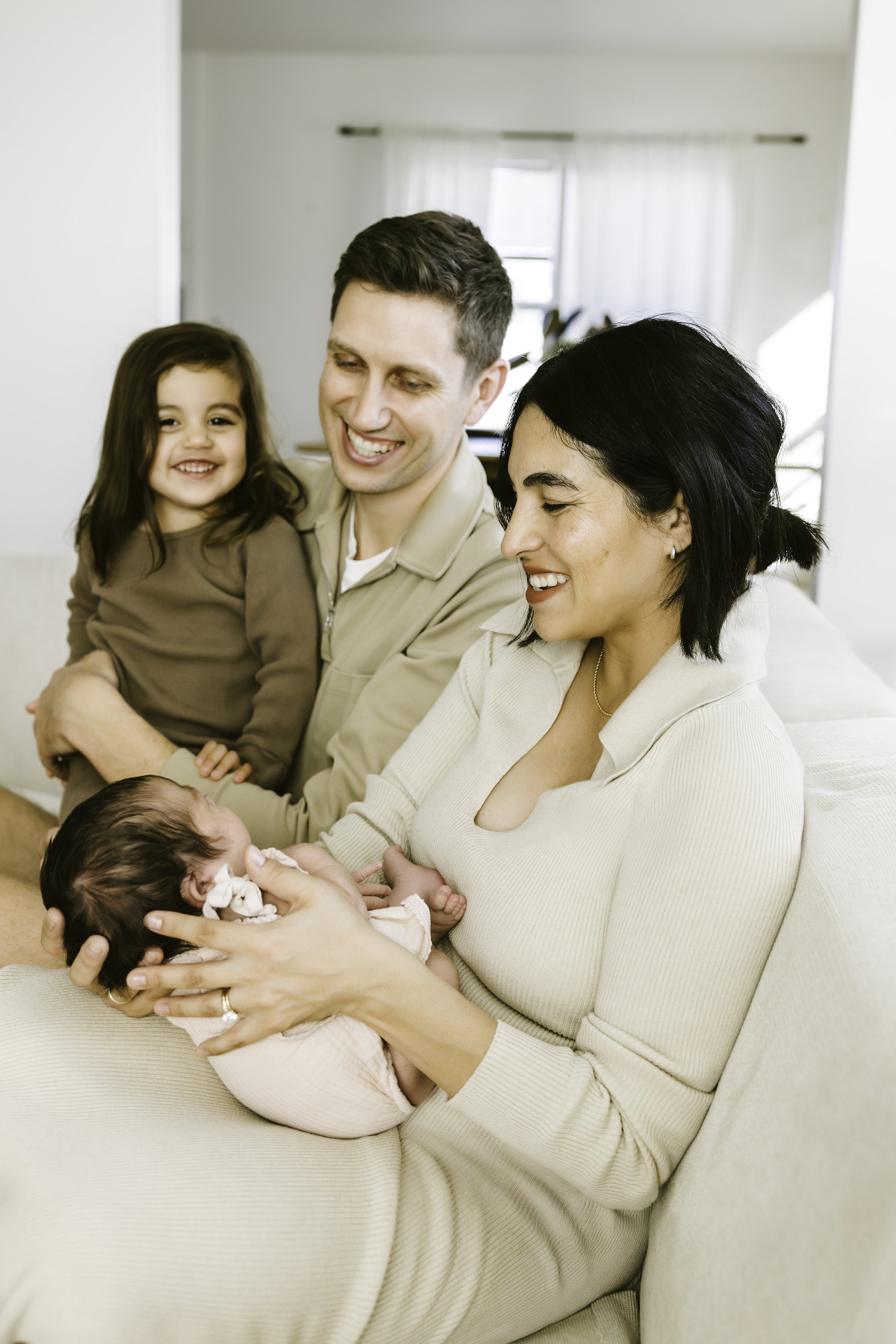 Family of four with a newborn, a woman holding the baby, a man and a young girl, all sitting on a white couch in a bright living room.