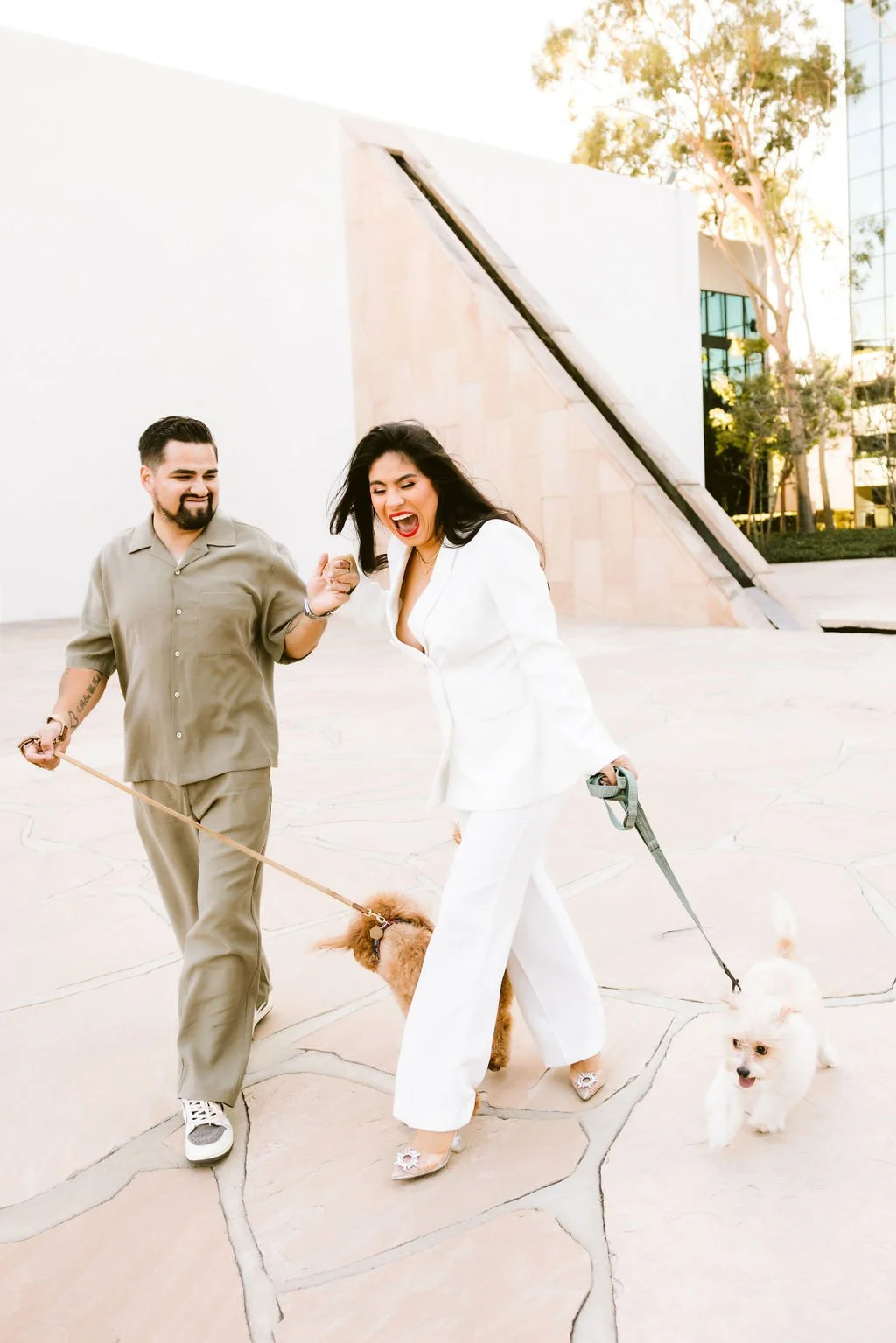 A man and woman laughing and holding leashes while walking dogs outdoors near a modern building and trees.