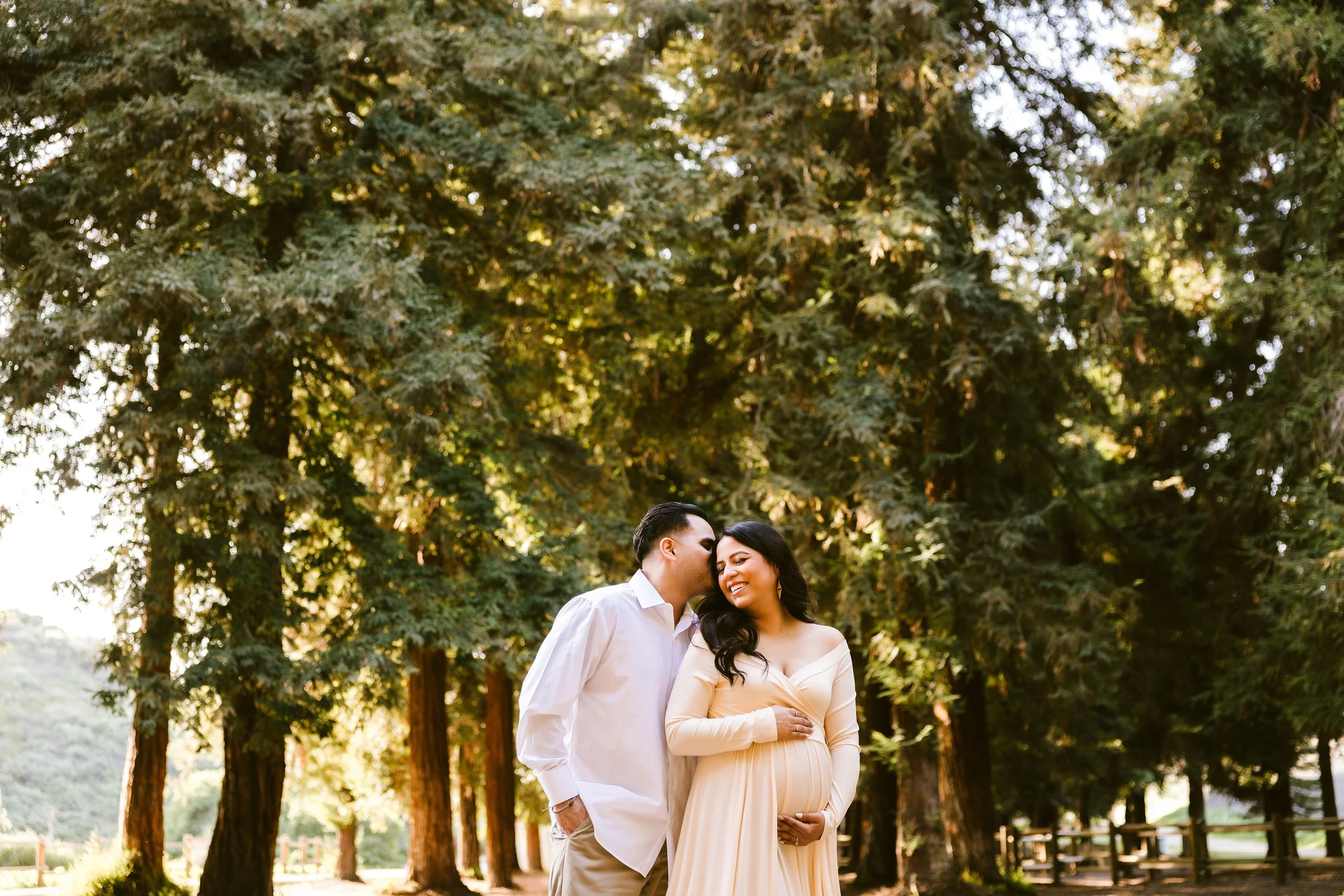 A smiling pregnant woman in a long beige dress with dark hair, standing under tall green trees, while a man in a white shirt and beige pants kisses her cheek, in a park setting.